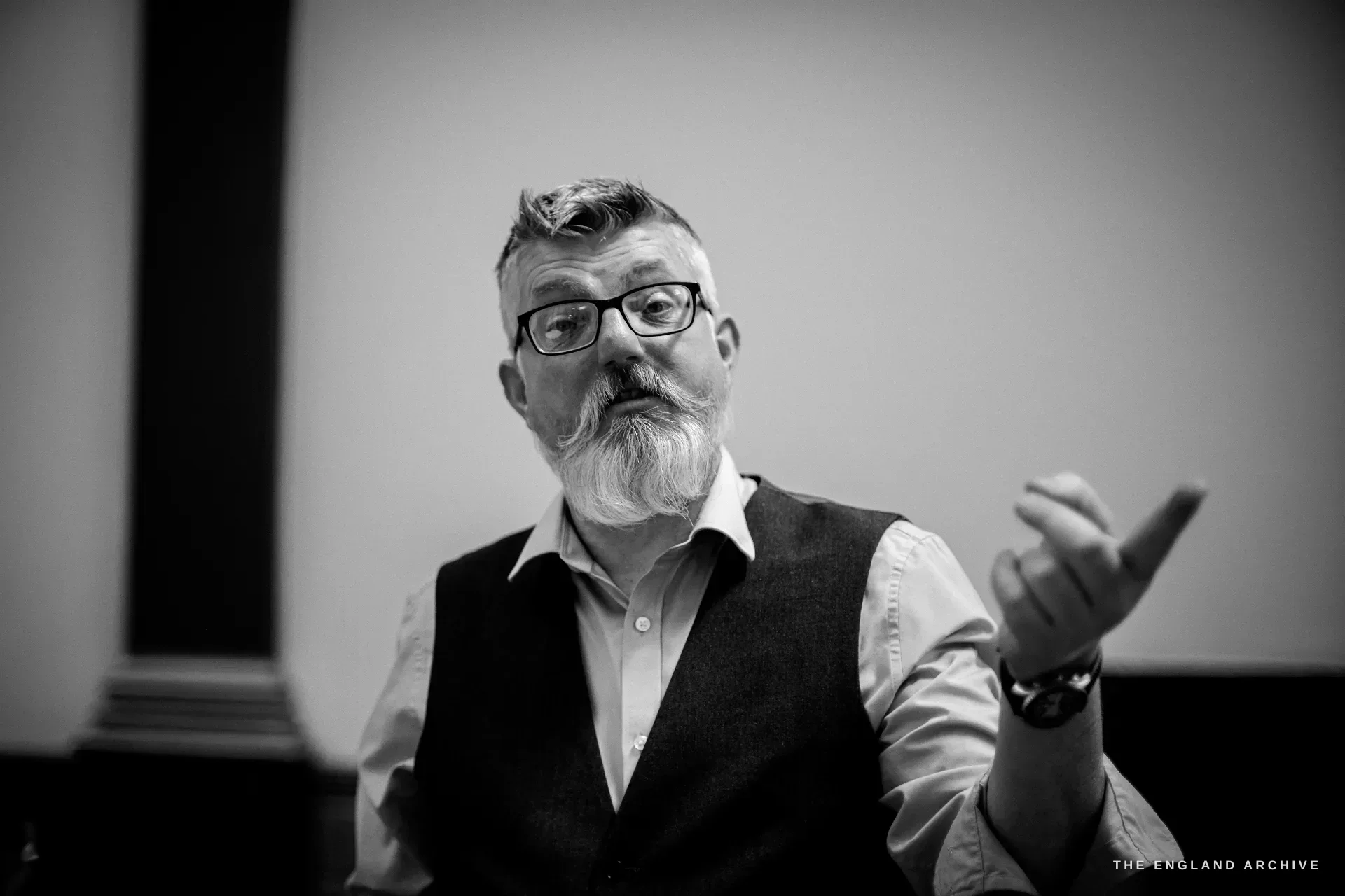 A bearded man in glasses and a waistcoat talking, one hand raised, a pale panelled wall behind him.