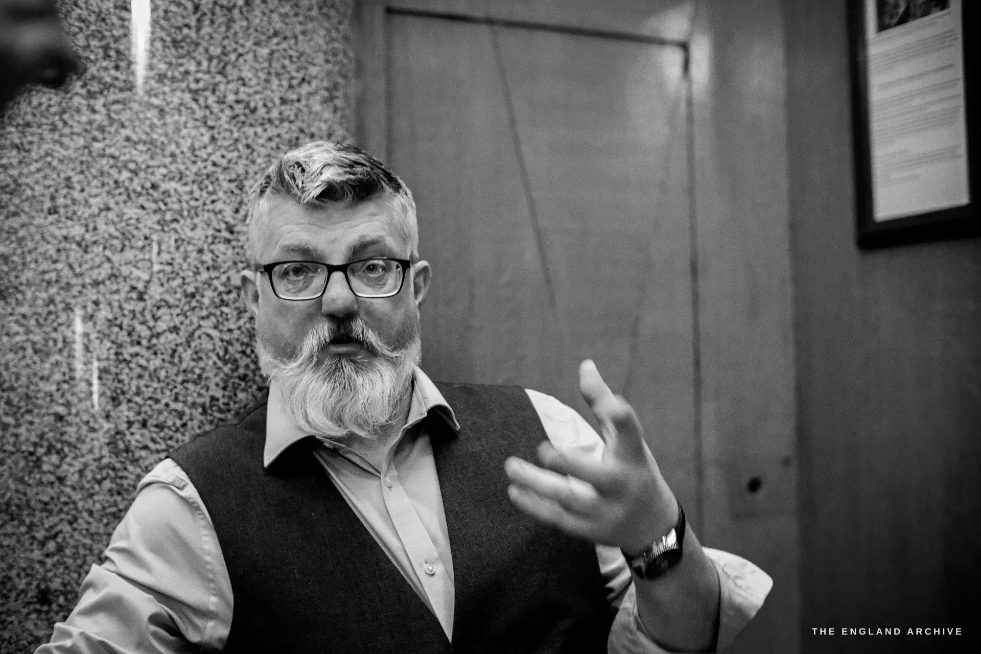A close portrait of a bearded man in glasses and a waistcoat, one hand raised, mid-sentence.