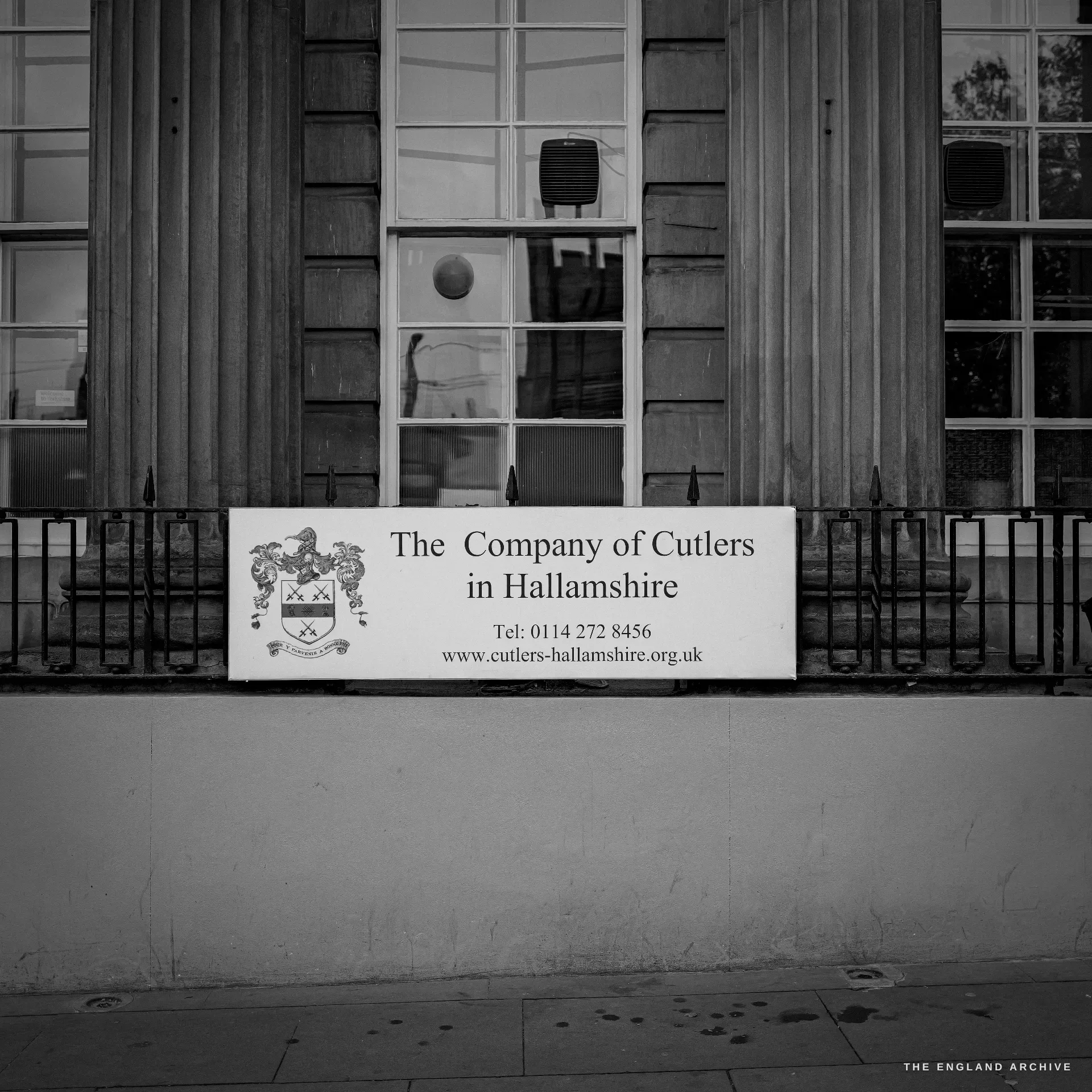 The classical stone frontage of Cutlers' Hall on Church Street, a sign reading 'The Company of Cutlers in Hallamshire' fixed to the railings below the columns.