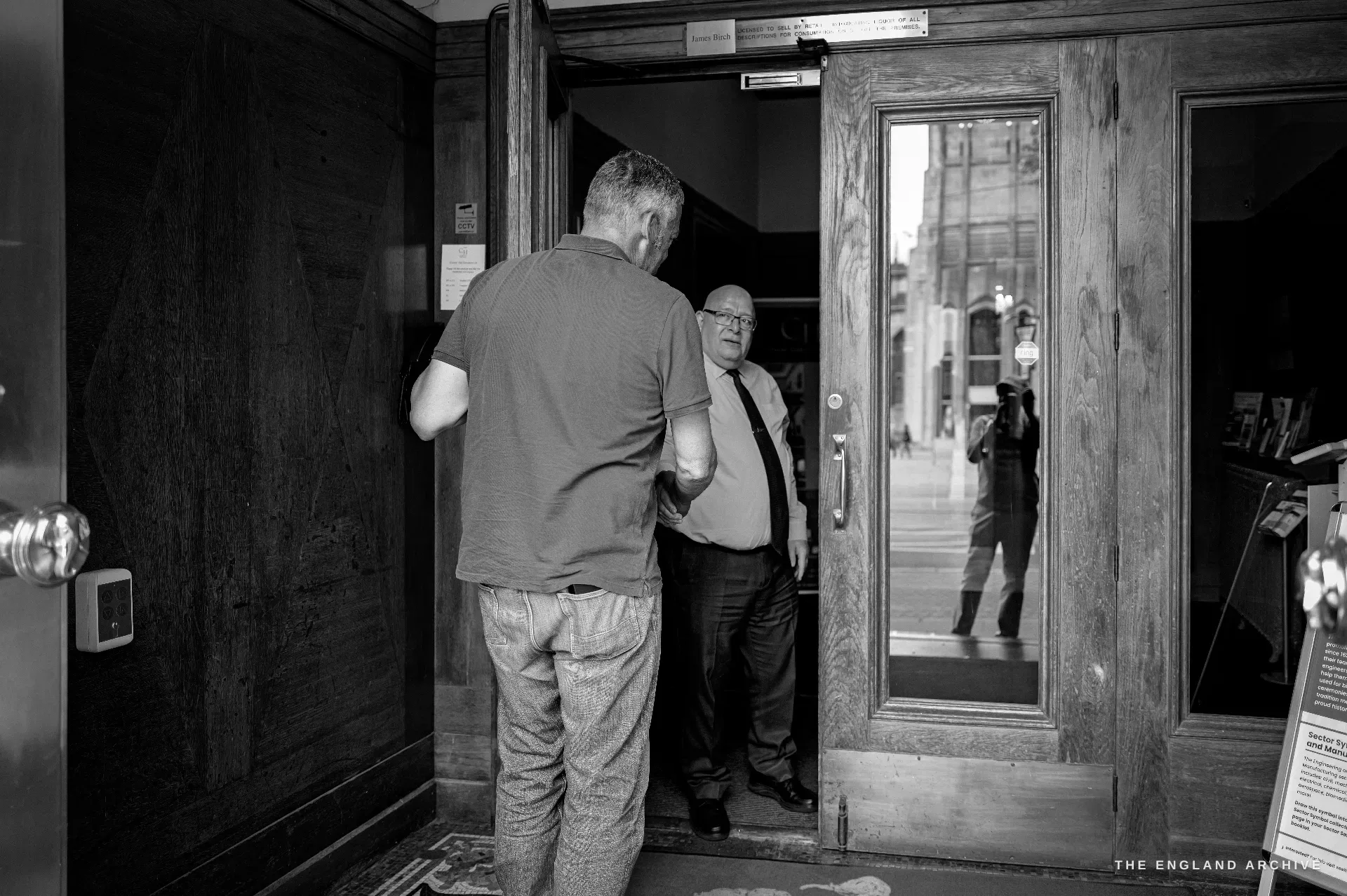 A visitor greeted at the open oak doors of Cutlers' Hall by a suited attendant, the street and the cathedral reflected in the glass of the door.