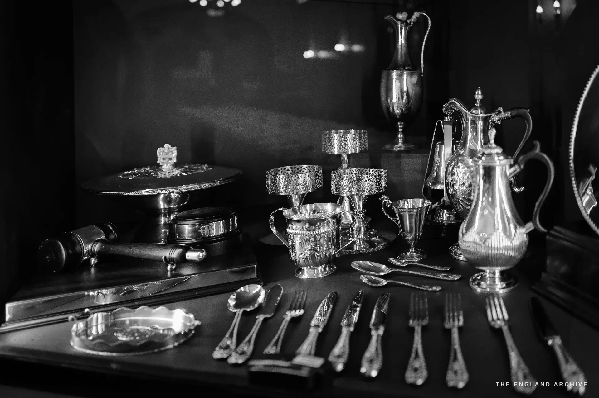 A close still-life of Company silver on green baize: coffee pots, a two-handled cup, a wine ewer, a wooden gavel, and cutlery with ornate handles.