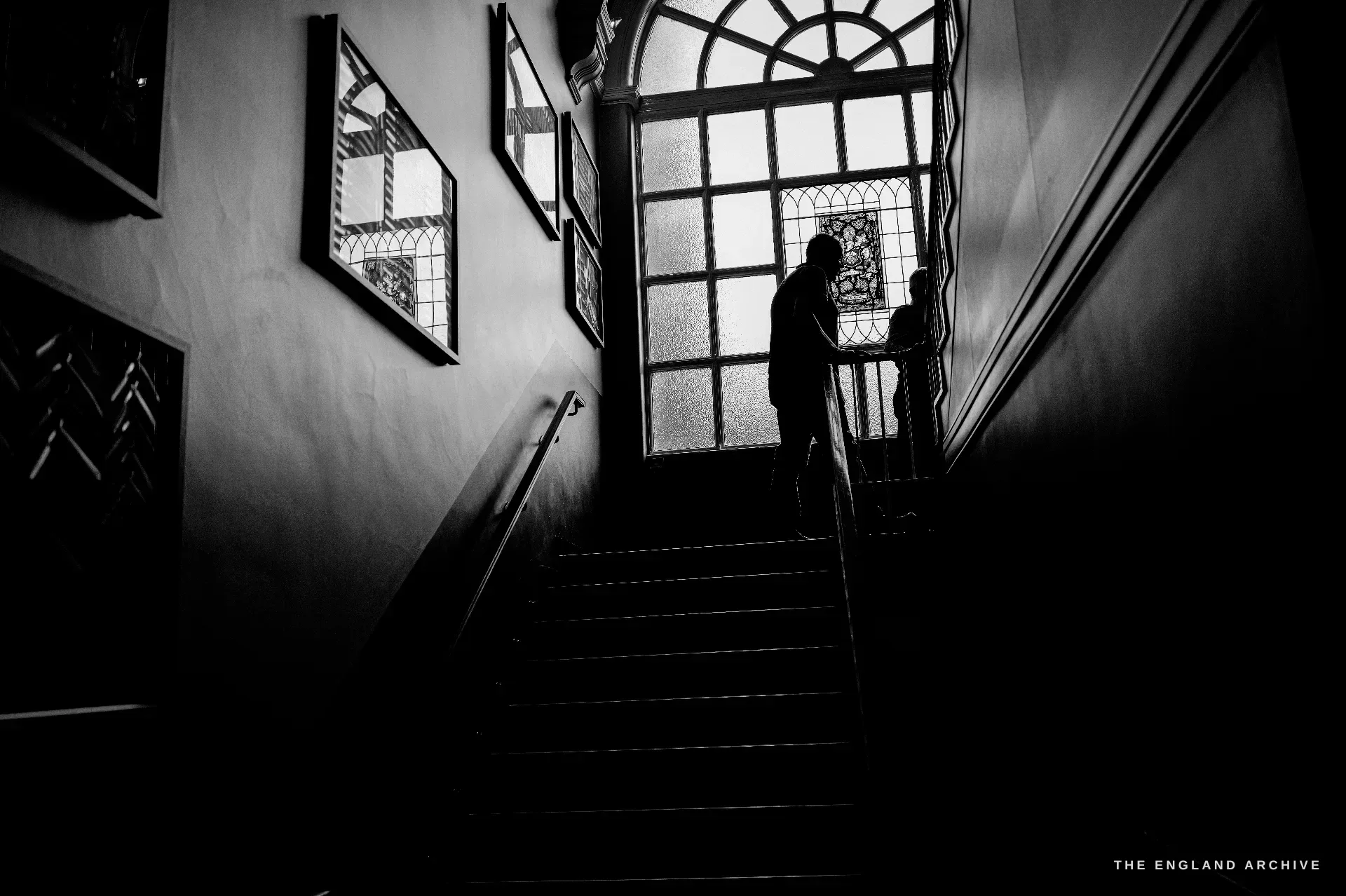 The Hall's staircase in silhouette against a tall arched window of leaded and stained glass, two figures climbing towards the light.