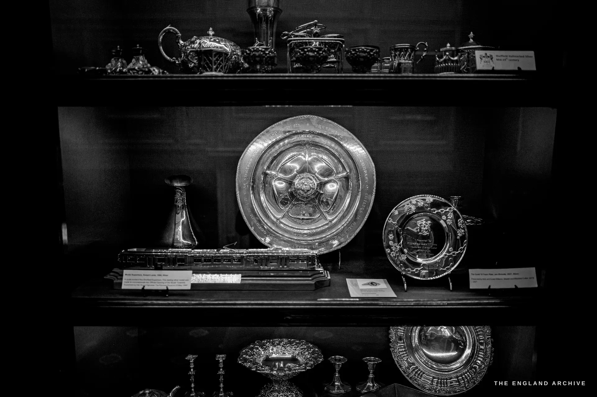 A display cabinet of Sheffield hallmarked silver: a large engraved presentation salver, a silver model of a Supertram, and smaller labelled pieces.