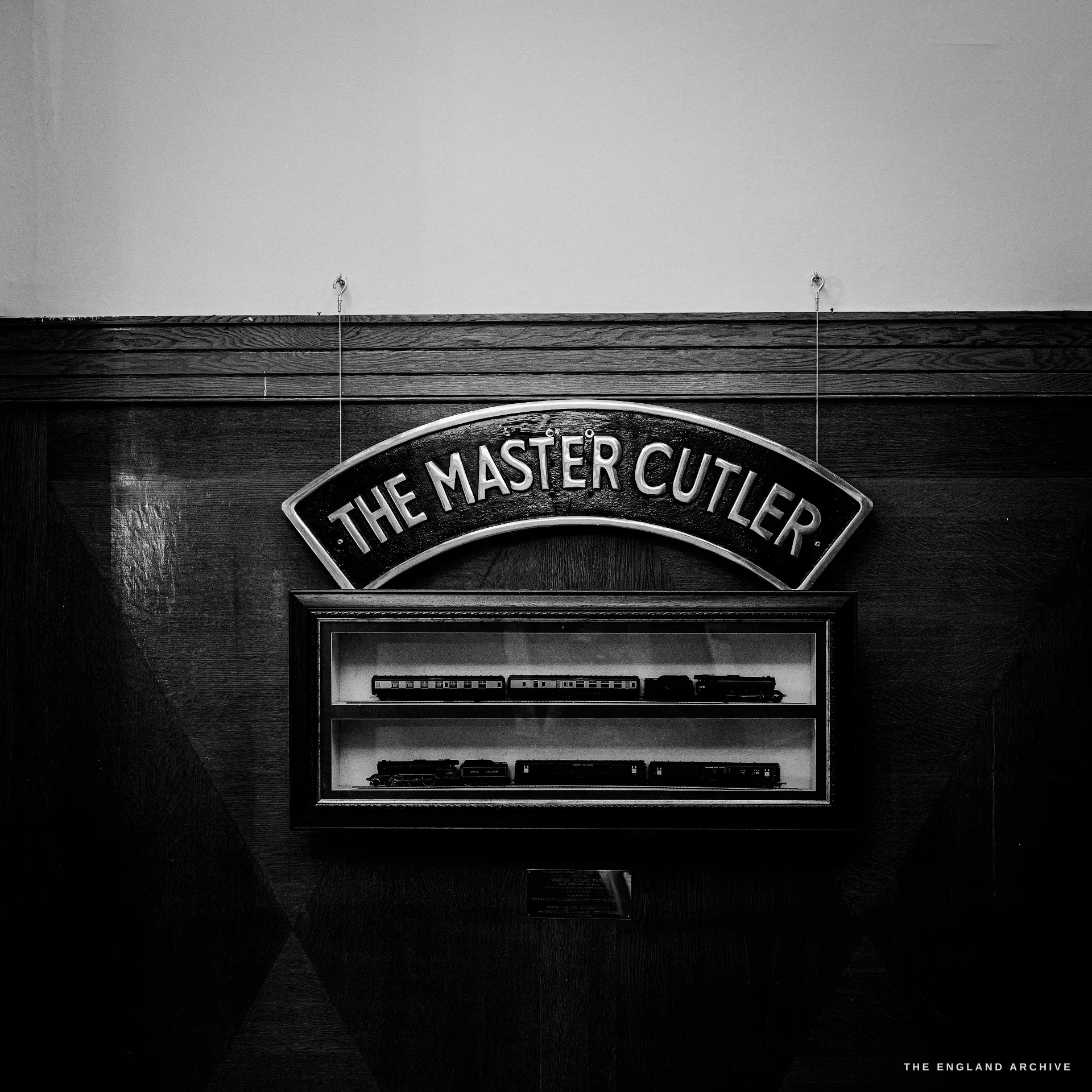 A cast railway nameplate reading 'THE MASTER CUTLER' mounted on oak panelling above a glazed case of model locomotives.