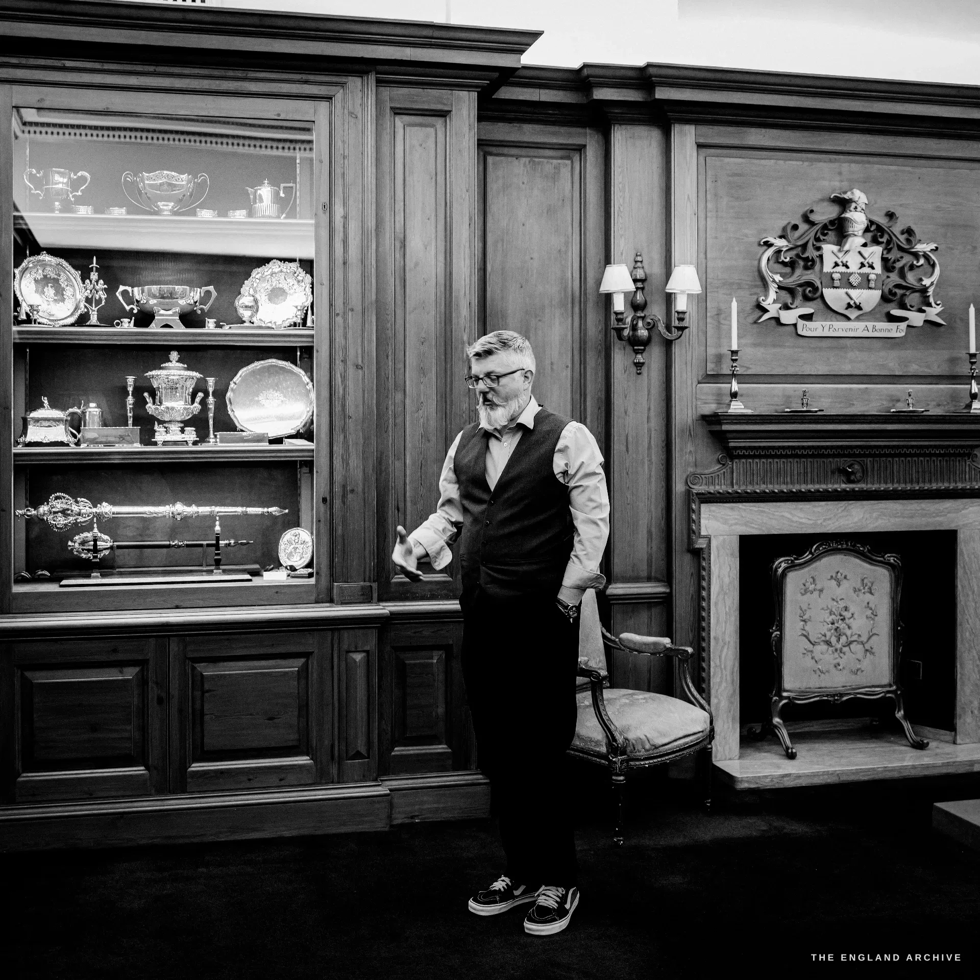 A man in a waistcoat gestures as he talks, standing between a lit cabinet of ceremonial silver, a mace among it, and a carved oak overmantel bearing the Company's arms and motto.