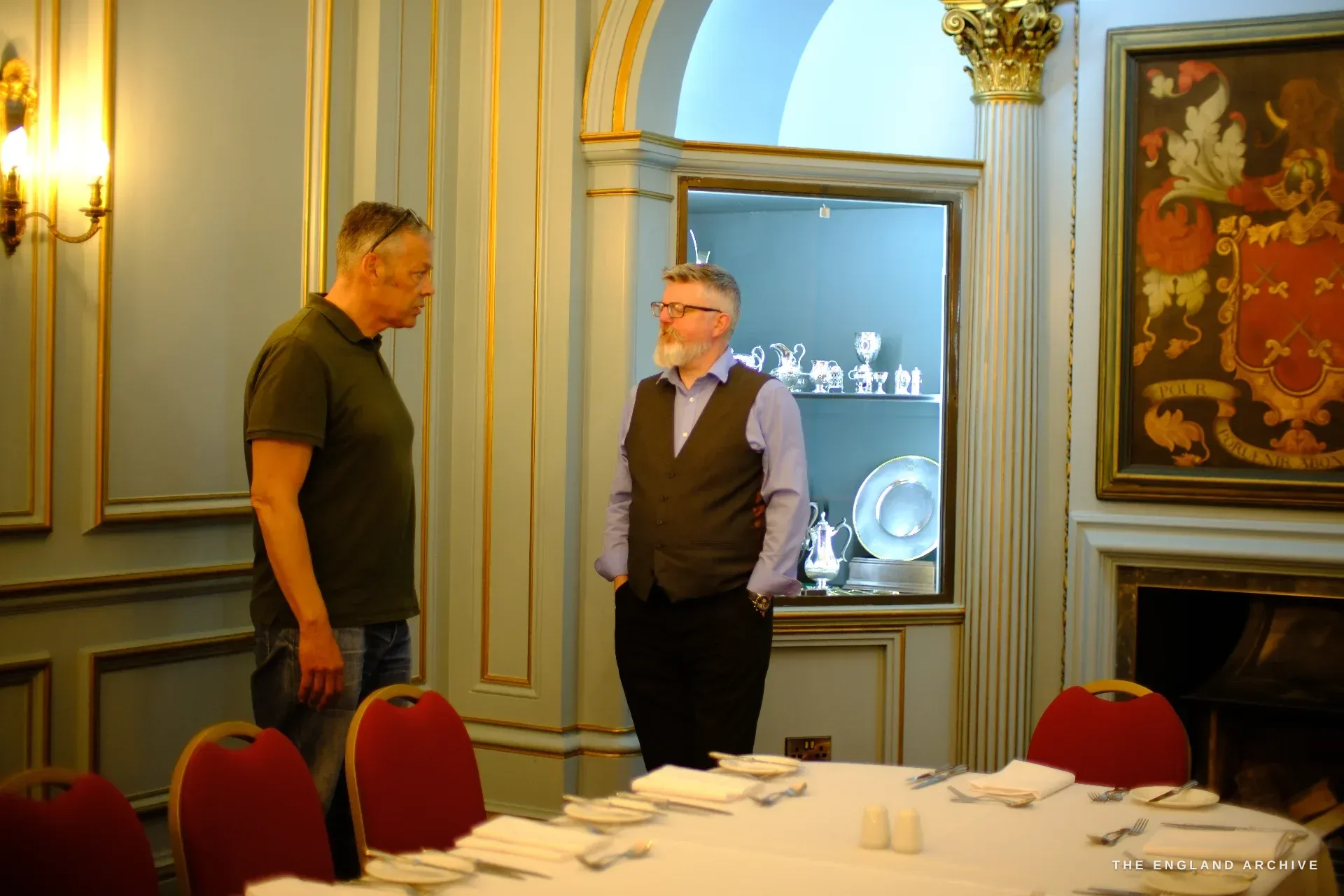 A blue-and-gold panelled dining room in Cutlers' Hall set for a meal, two men in conversation beside a niche of silver, the painted Company arms on the wall.