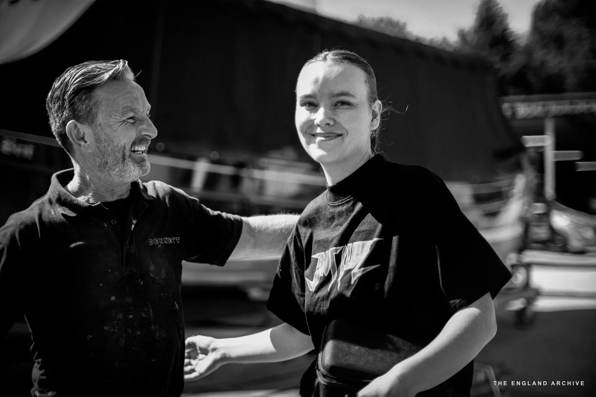 Stephen Dennett (left, in DENNETT polo, smiling) standing in the yard with Lucy, a younger collaborator (right, in dark T-shirt, smiling toward Stephen). Boats and the workshop tarpaulin behind them.