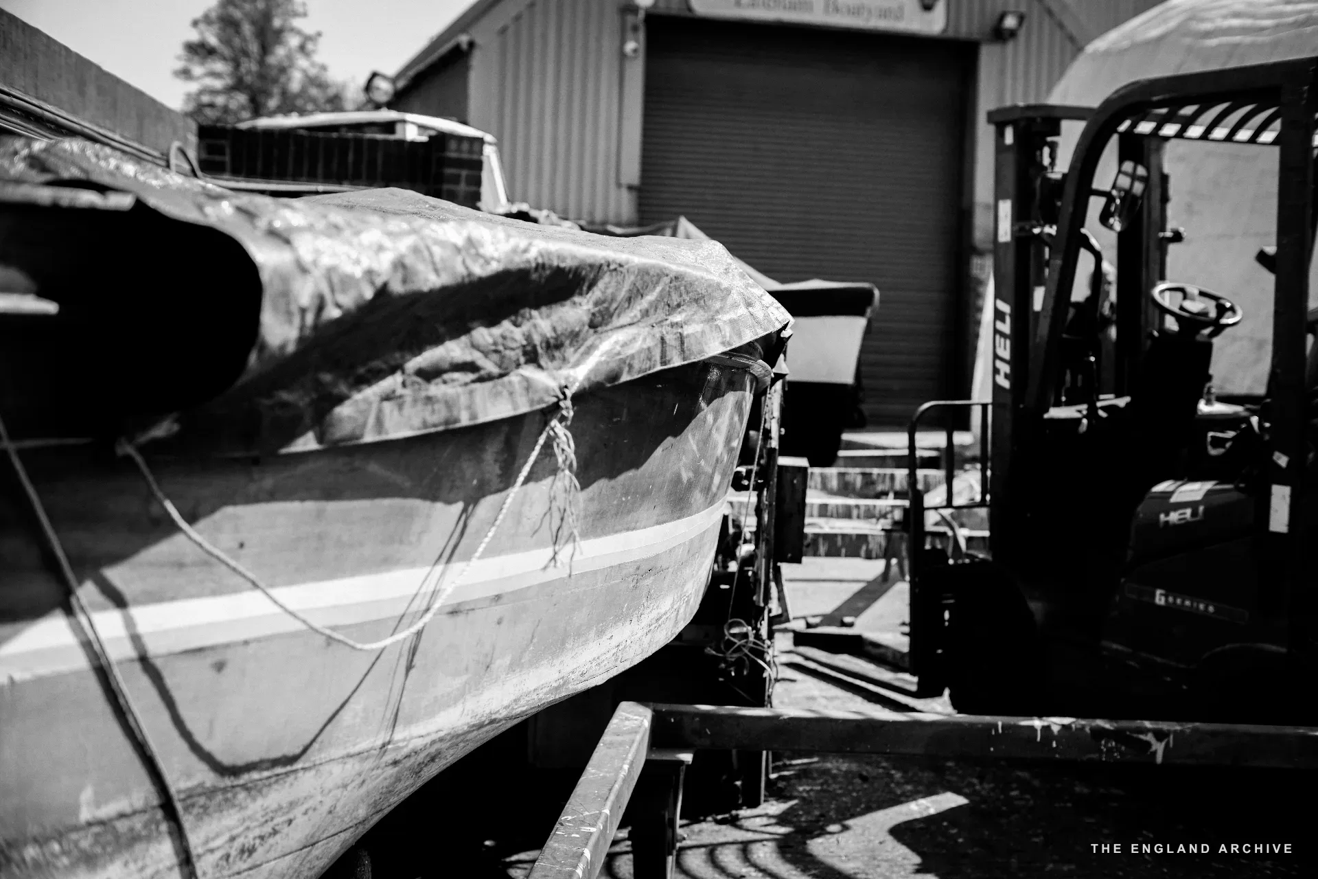 A wooden boat under tarpaulin in the yard, propped on a trailer, with a forklift parked alongside and the workshop building behind.