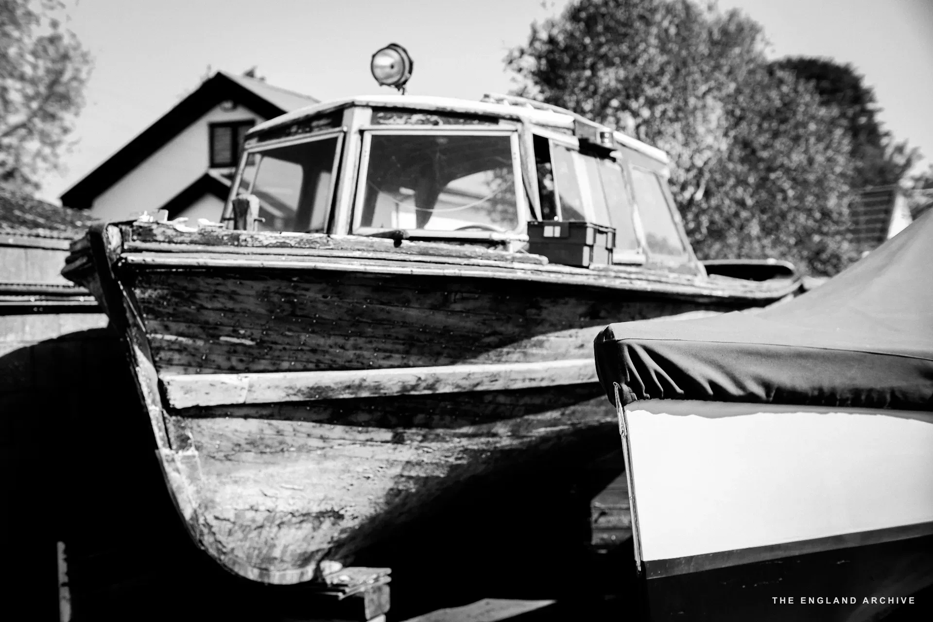 The bow of a wooden cabin cruiser parked in the yard, varnished hull catching the light, the workshop’s pitched roof behind.