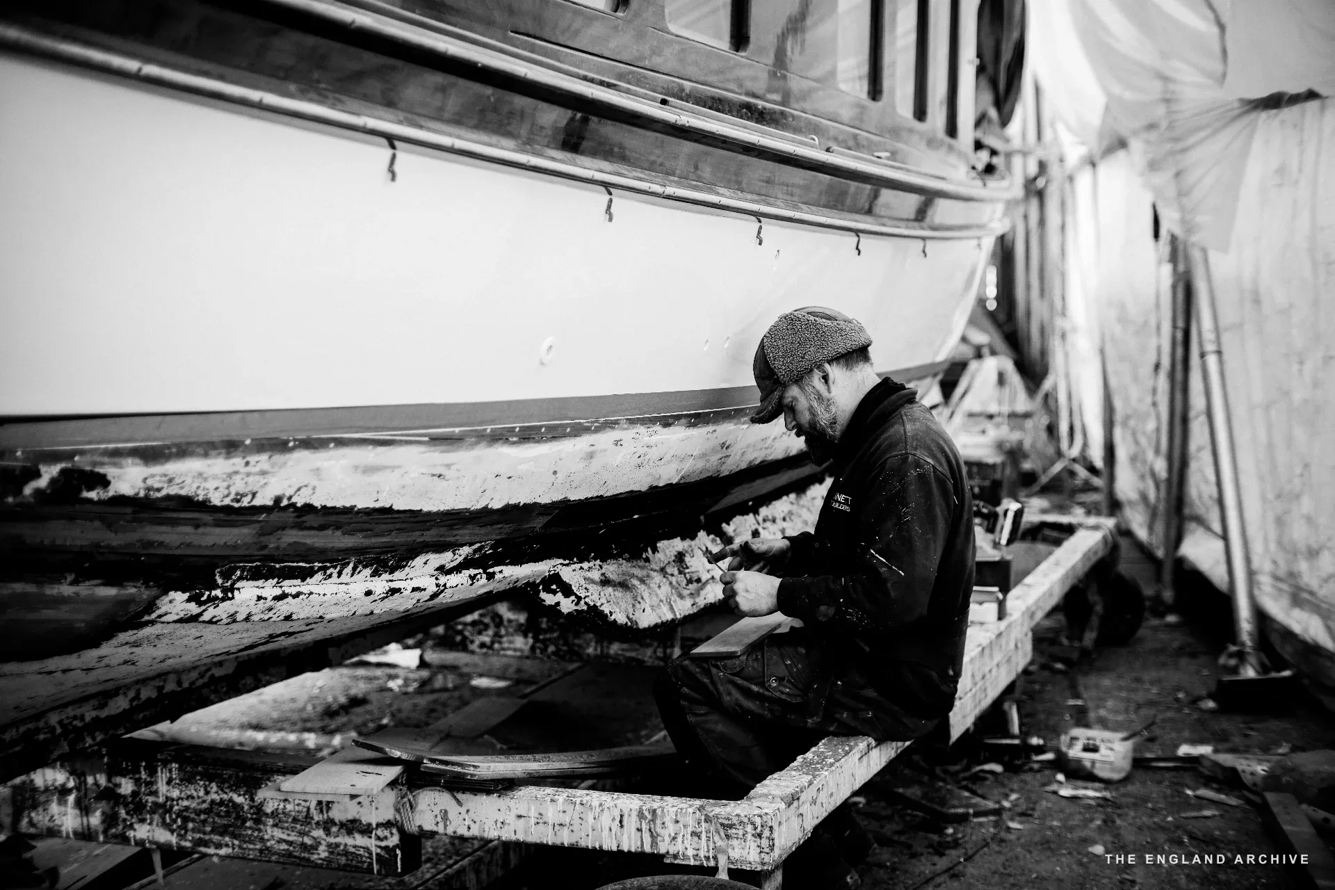 A worker in a flat cap kneeling beside the keel of a wooden boat in the workshop, working at the boat’s underside on the trestles.
