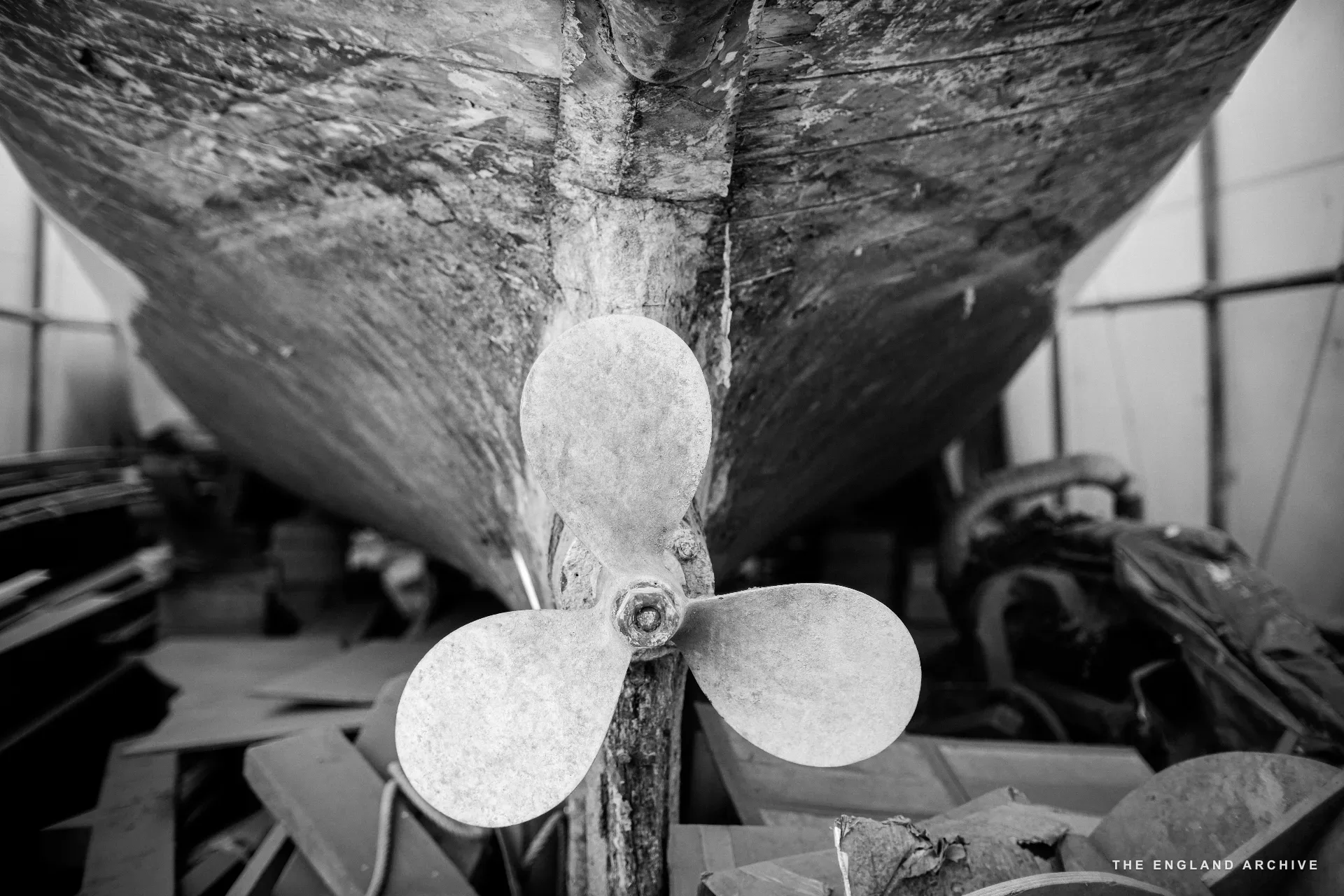 The propeller and rudder of a wooden hull seen from the bottom, the keel rising overhead in the workshop.