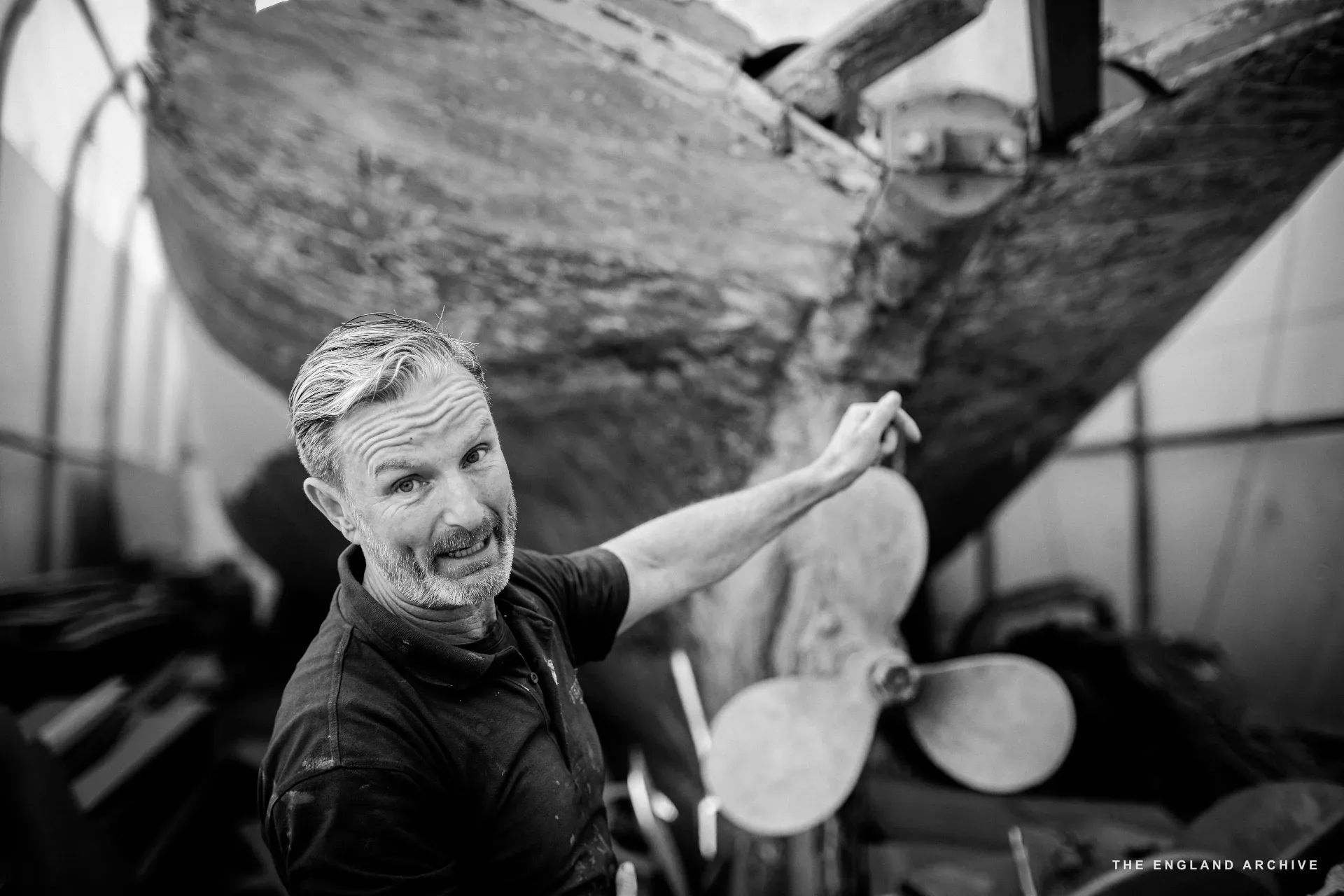 Stephen Dennett standing beside the propeller of a wooden hull in the workshop, looking toward the camera, hand resting on the timber above the prop.