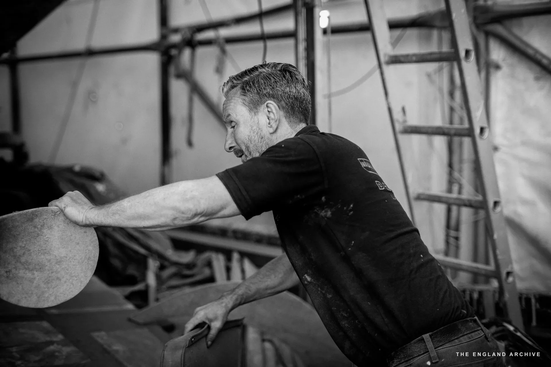 Stephen Dennett in motion at the workshop, holding a circular sanding pad mid-gesture, a ladder and the side of a hull behind him.