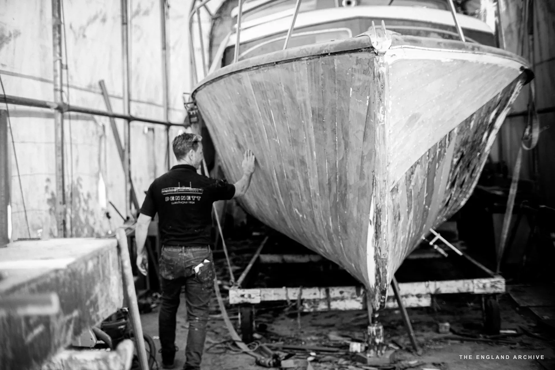 Stephen Dennett (back to camera, DENNETT polo visible) examining the bow of a wooden boat in the workshop, the timber bracing of the cradle visible below.