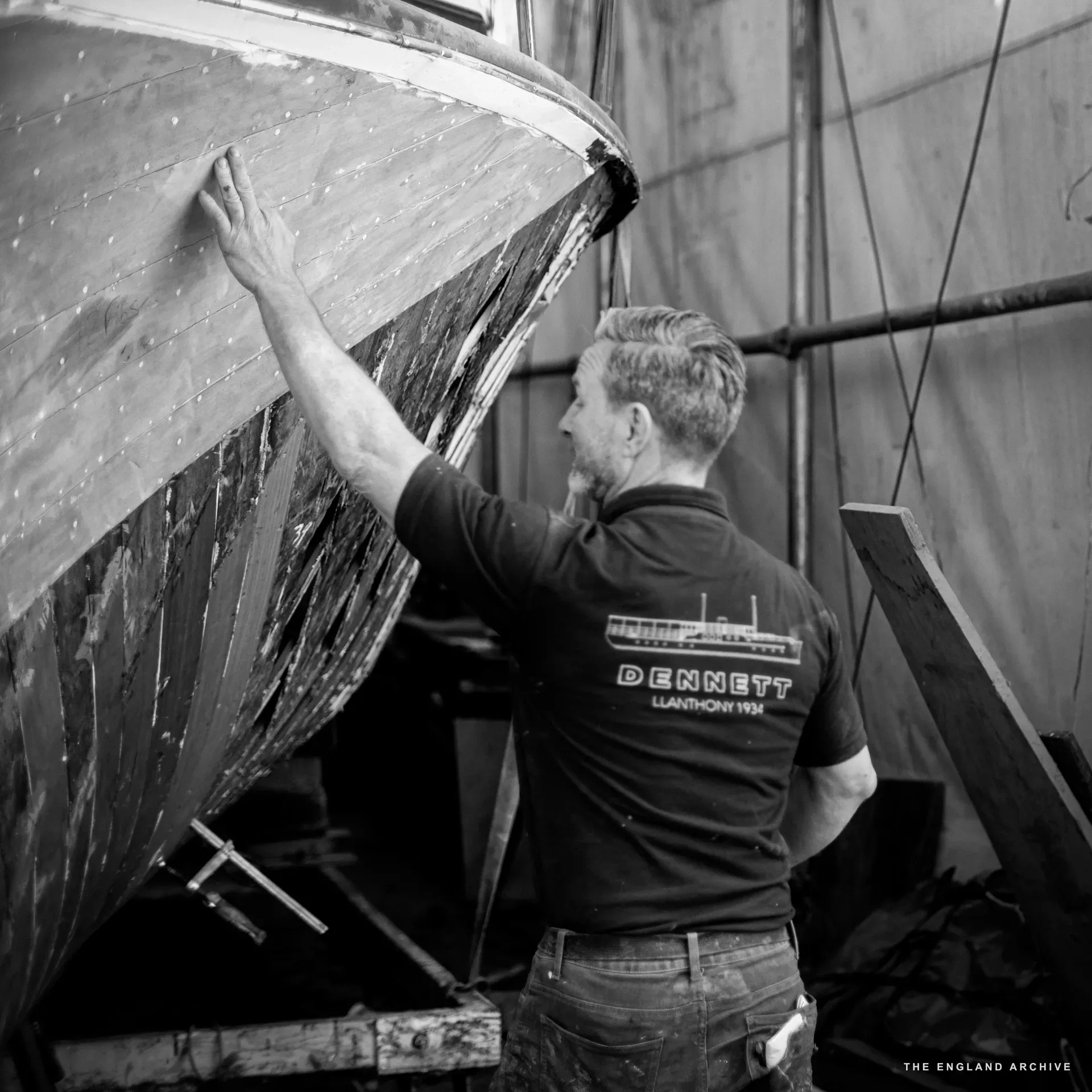 Stephen Dennett (back to camera, DENNETT LLANTHONY 1934 polo visible) running his hand along the side of a hull under restoration, examining the surface for fairness.
