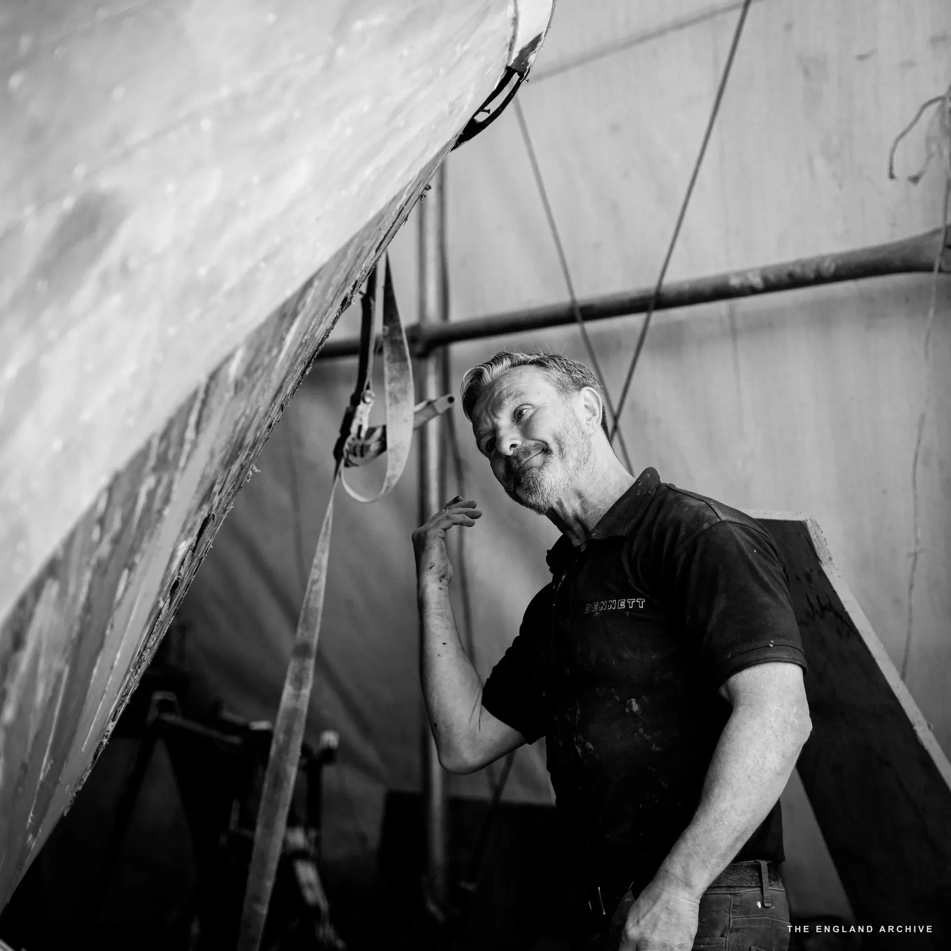 Stephen Dennett looking up at the bow of a large wooden hull, faint smile, hand resting on the underside of the boat. Workshop light from the right.