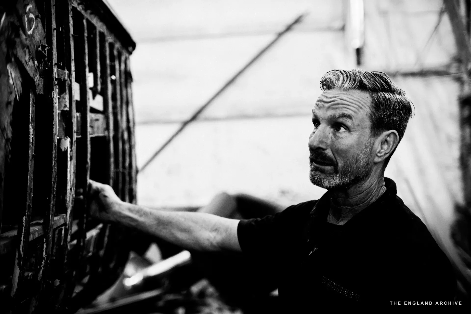 Stephen Dennett at a metal bench grate in the workshop, hand on the grate, looking up and away.