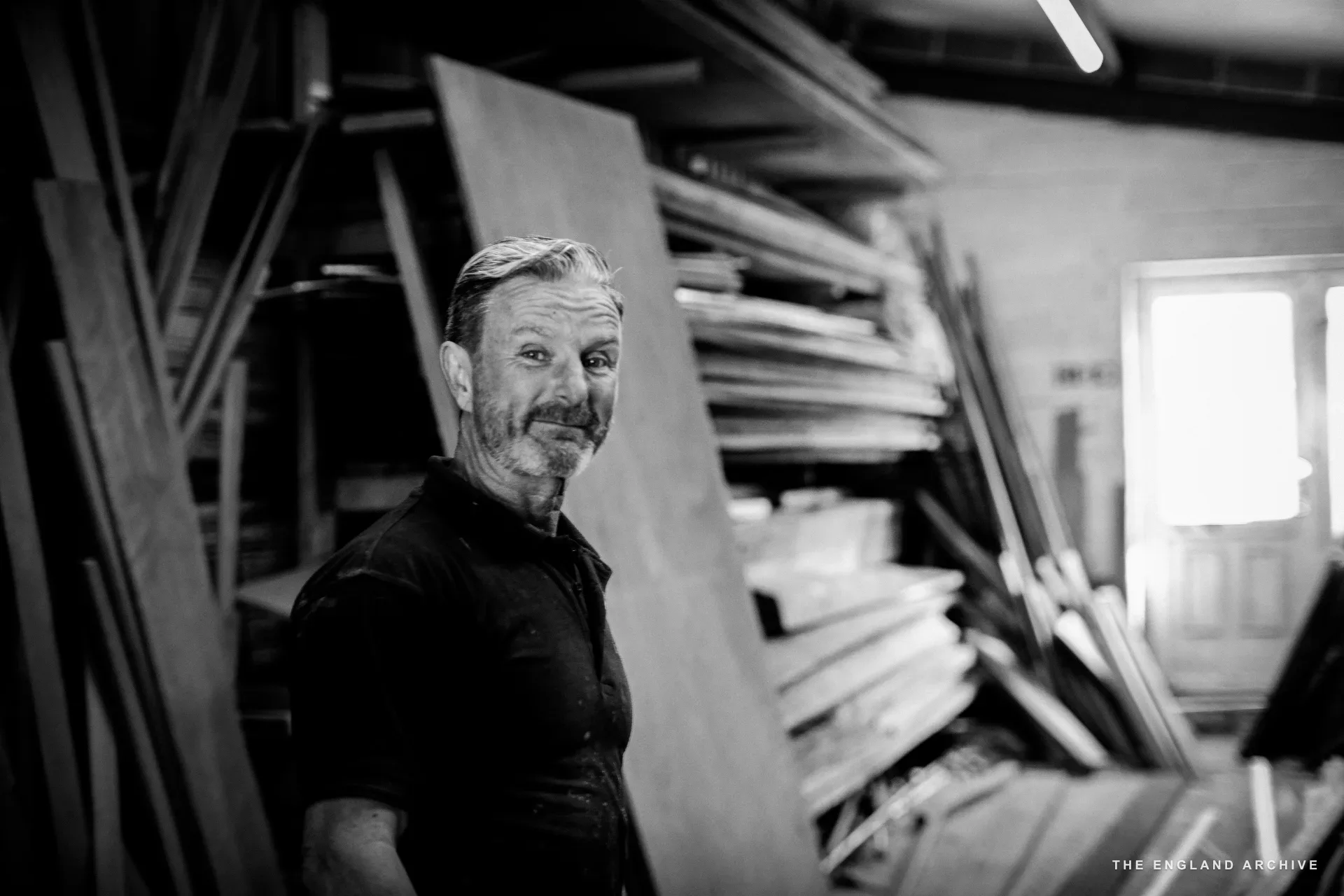 Stephen Dennett standing in the timber-store mezzanine of the workshop, a tall stack of plywood sheets and shelves of long timbers behind him, looking at the camera.