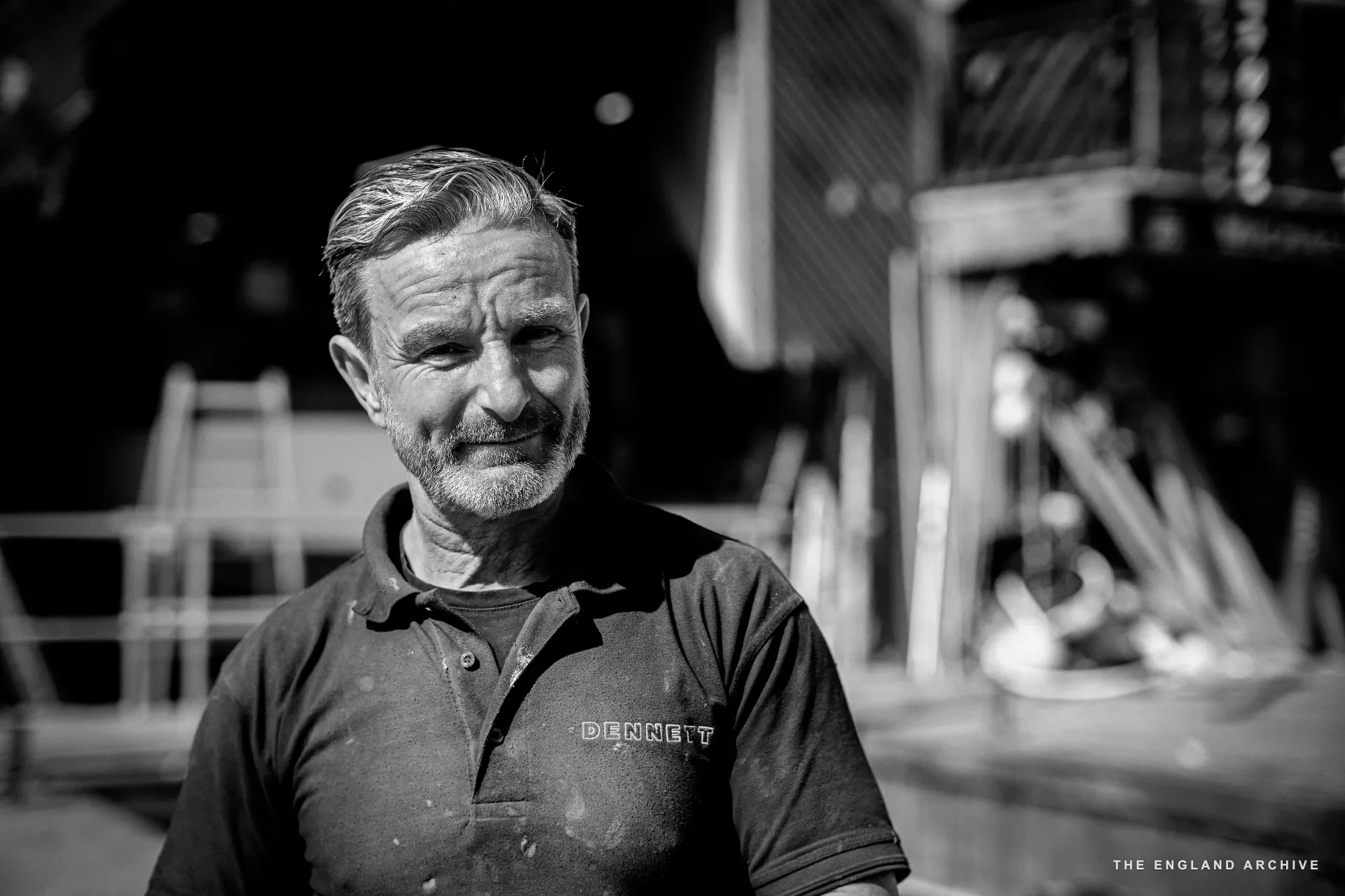 A head-and-shoulders portrait of Stephen Dennett in the workshop, looking down and slightly away with a faint smile, the DENNETT polo and a boat hull visible in the background.