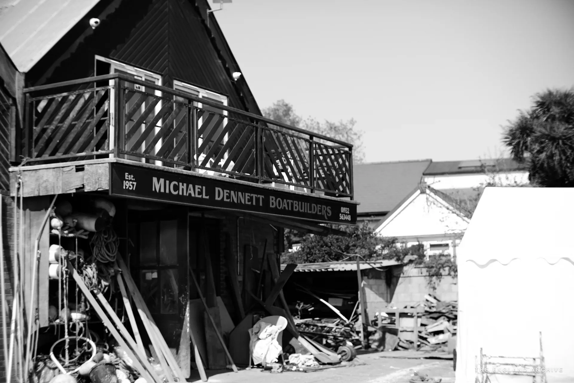 The exterior of the Dennett Boat Builders yard - the timber-clad workshop with the ‘Michael Dennett Boatbuilders Est. 1957’ sign mounted across its front, a balcony above and stacked materials below.