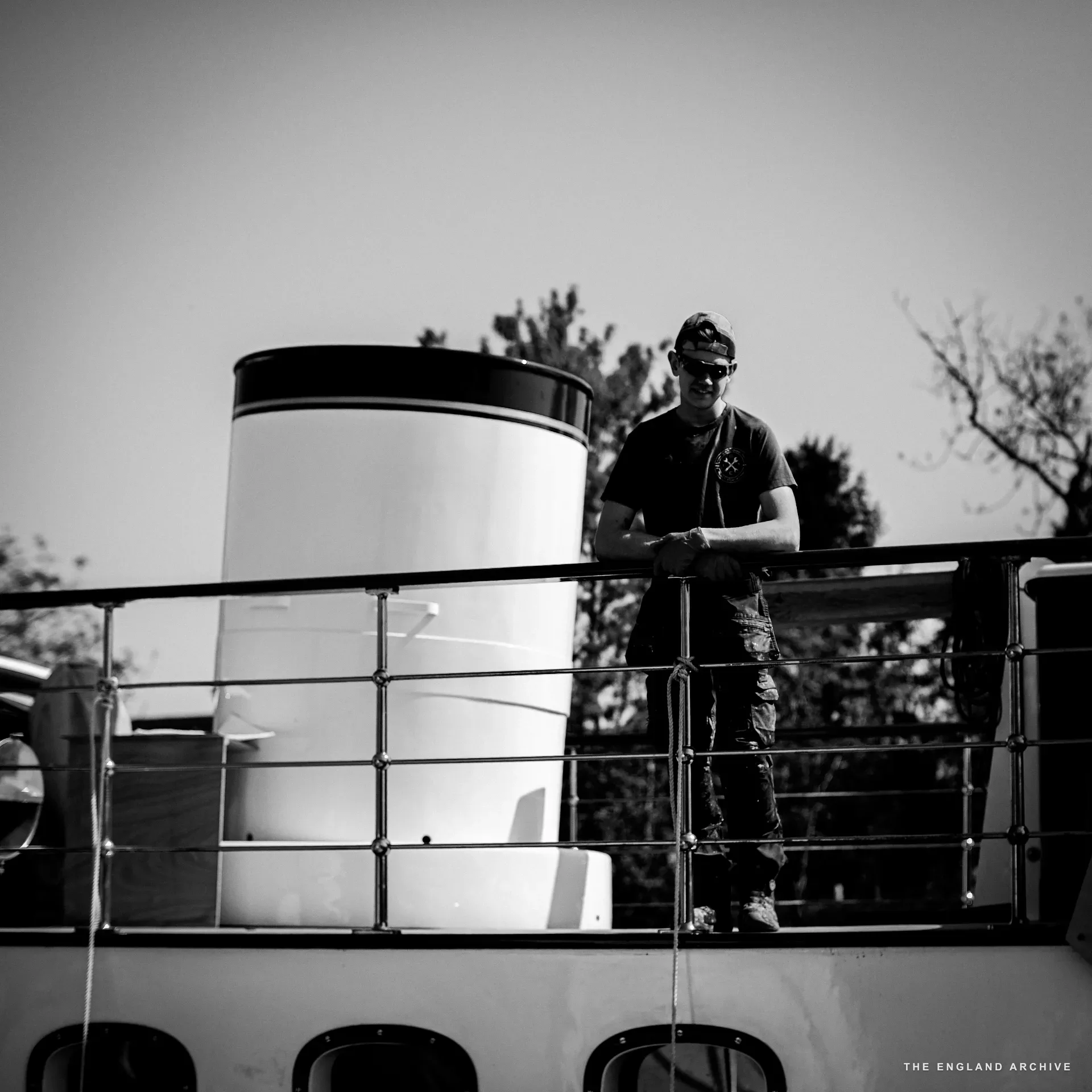 A worker in a flat cap and dark sunglasses standing on the upper deck of a yacht in the yard, hands on the safety rail, the white funnel beside him.