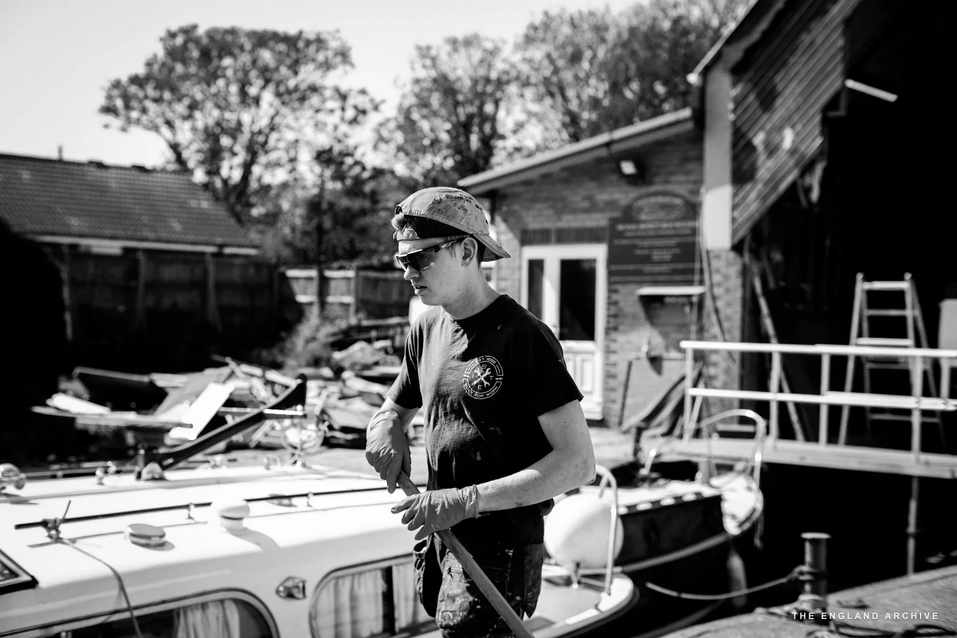 A worker in cap and sunglasses standing on the deck of a moored boat at the yard’s slipway, holding a hose, a wooden building and other moored boats behind.