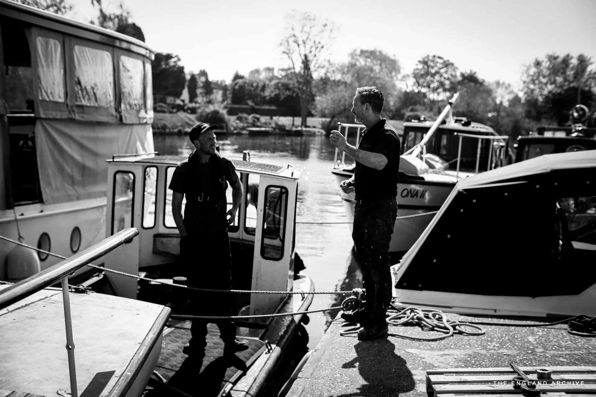 Two workers in conversation on the slipway between two boats, the river behind them with another moored vessel and willow-lined far bank.