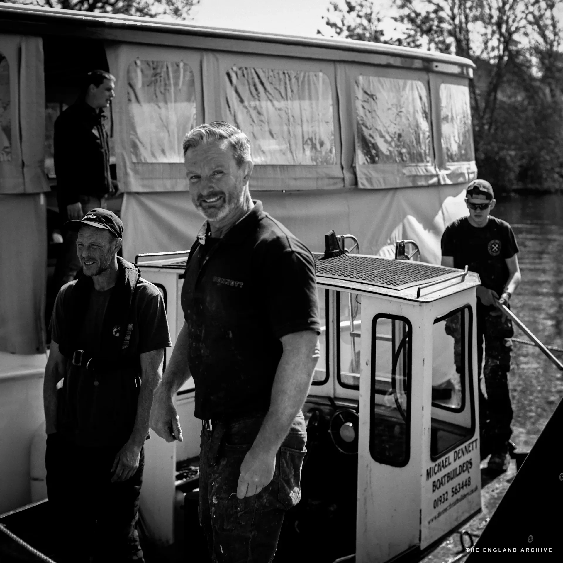 Stephen Dennett (centre, smiling) on the deck of a Dennett-built boat at the slipway with three other workers behind him, the yard’s phone number painted on the cabin side: ‘MICHAEL DENNETT BOATBUILDERS / 01932 563446’.