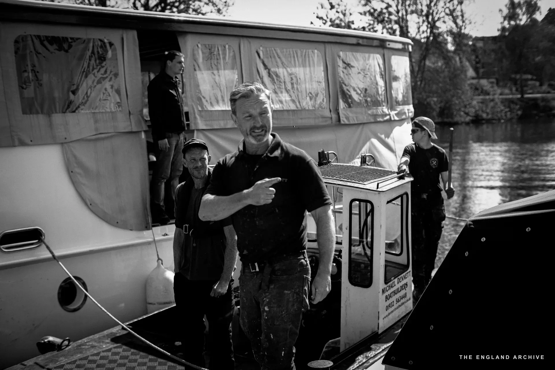Stephen Dennett pointing across the deck of the Dennett boat at the slipway, mid-gesture and laughing, with two other workers behind him by the cabin.