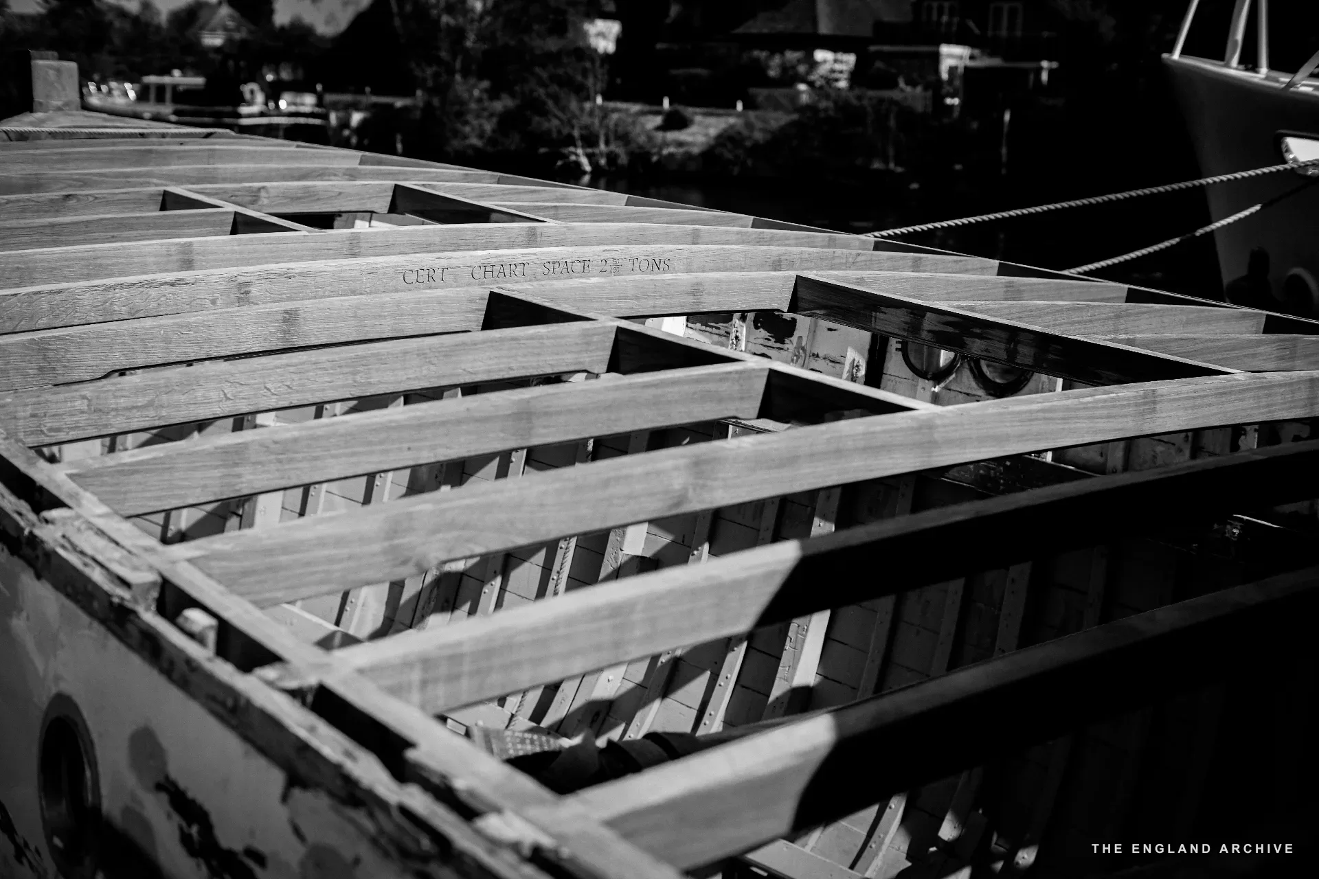 A boat hull seen from above with its deck framing exposed - timber ribs and stringers laid across the bottom of the hull, a stencilled inscription faintly visible on one rib.