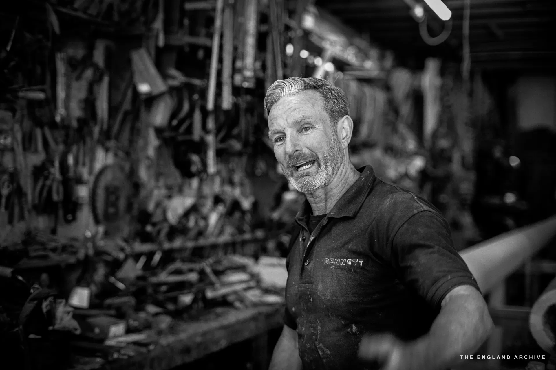 Stephen Dennett standing in front of the workshop’s tool wall, head turned, broad smile, the dense rack of hand tools behind him.