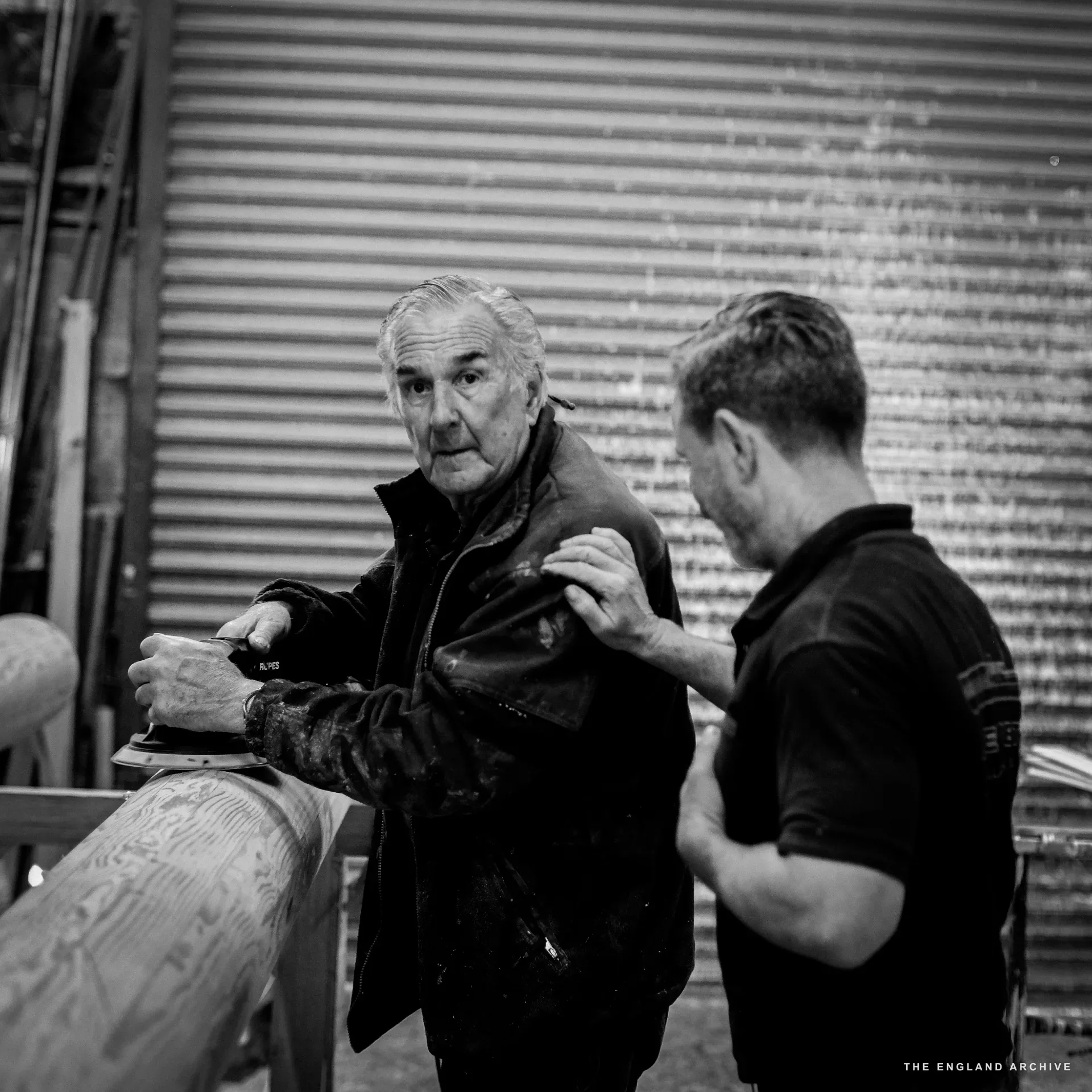 Michael Dennett (left, white hair, jacket) and Stephen Dennett (right, in DENNETT polo) at a workbench together - Michael working a rotary sander on a long timber, Stephen standing close by with a hand on Michael’s shoulder. The corrugated workshop door behind.