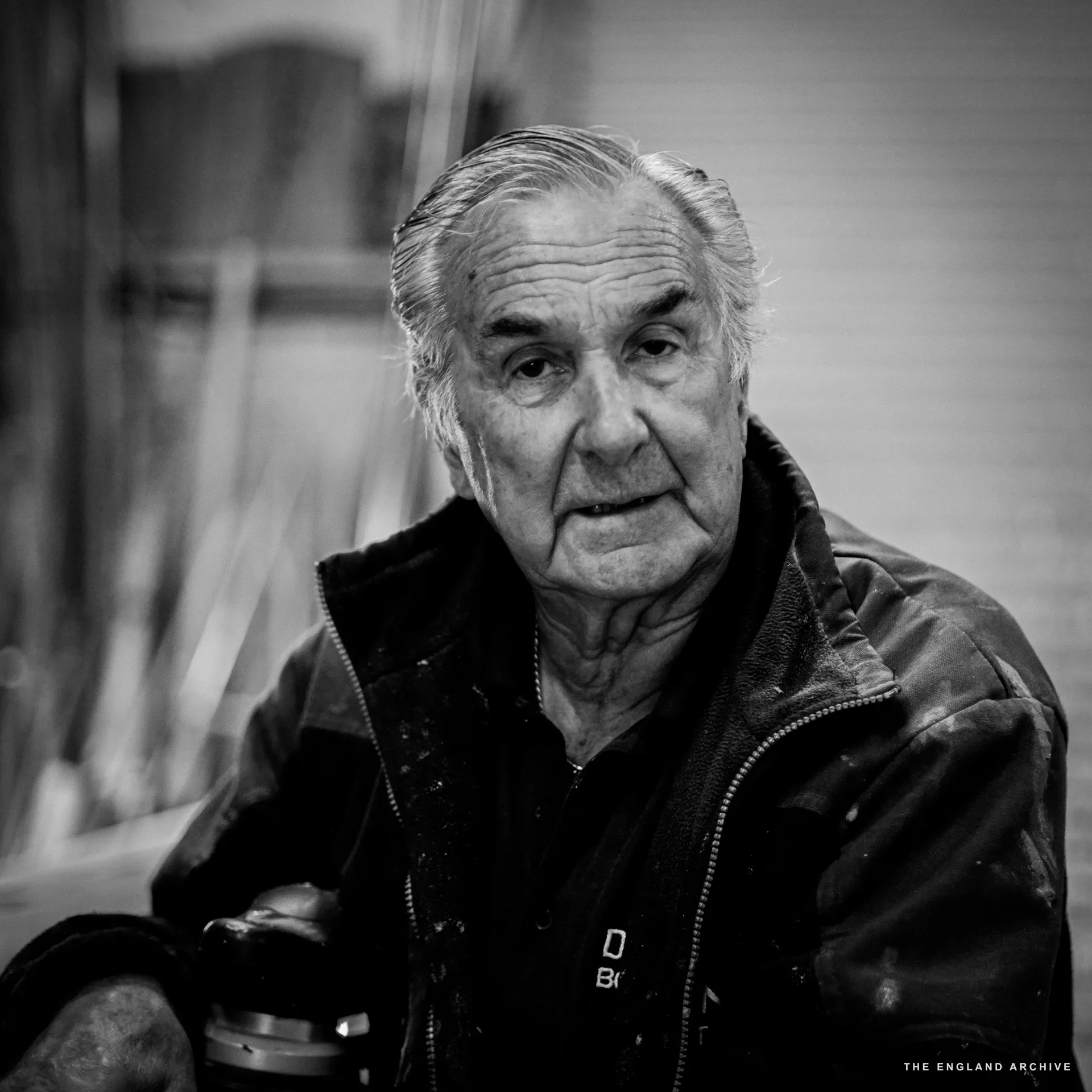 A close head-and-shoulders portrait of Michael Dennett, white hair, looking directly at the camera, dark jacket worn over the DENNETT polo, the workshop softly out of focus behind.