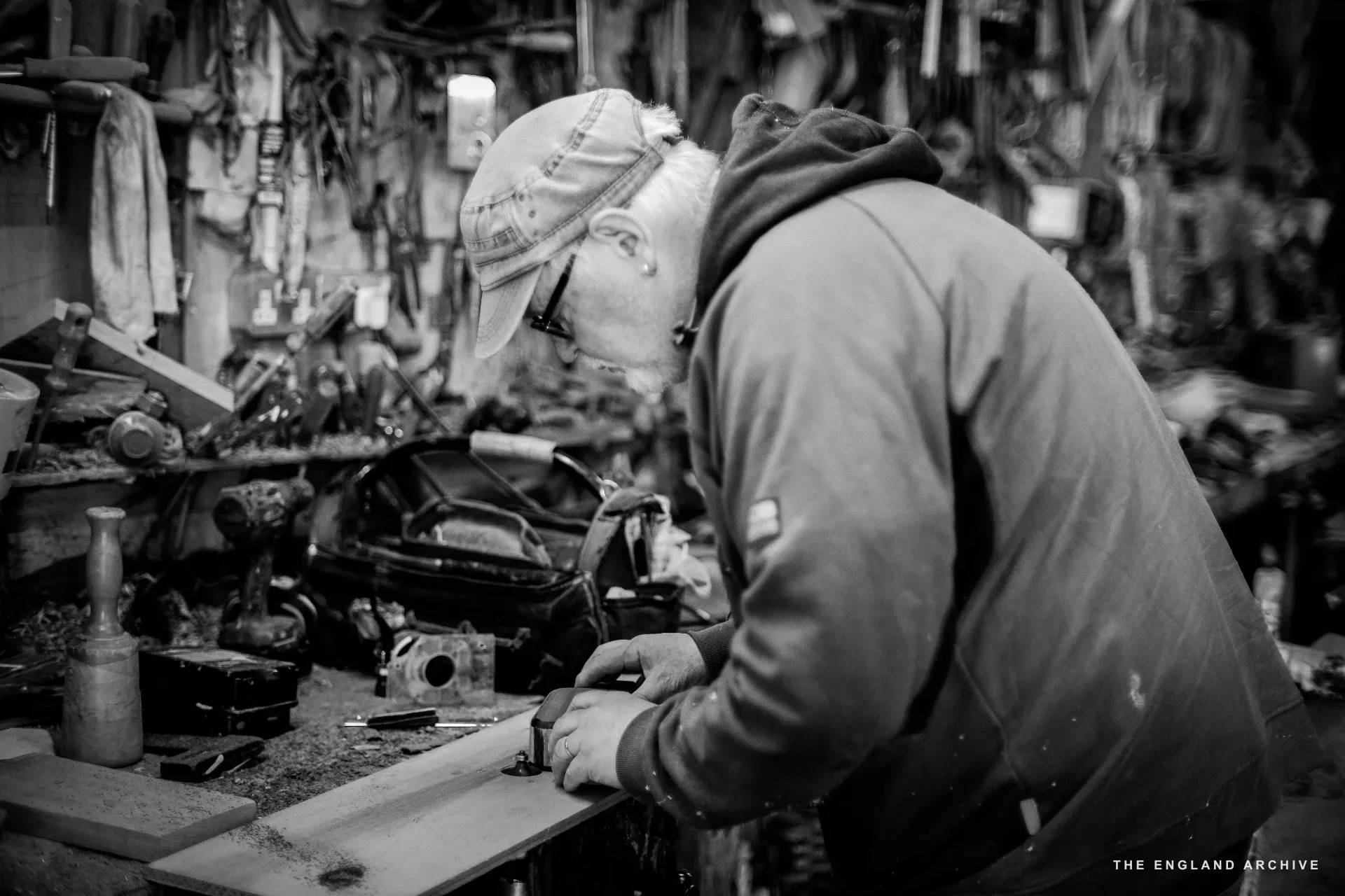 A worker in a flat cap and grey sweatshirt working at the cluttered tool wall bench, head down on the work, the whole tool rack rising above the bench.
