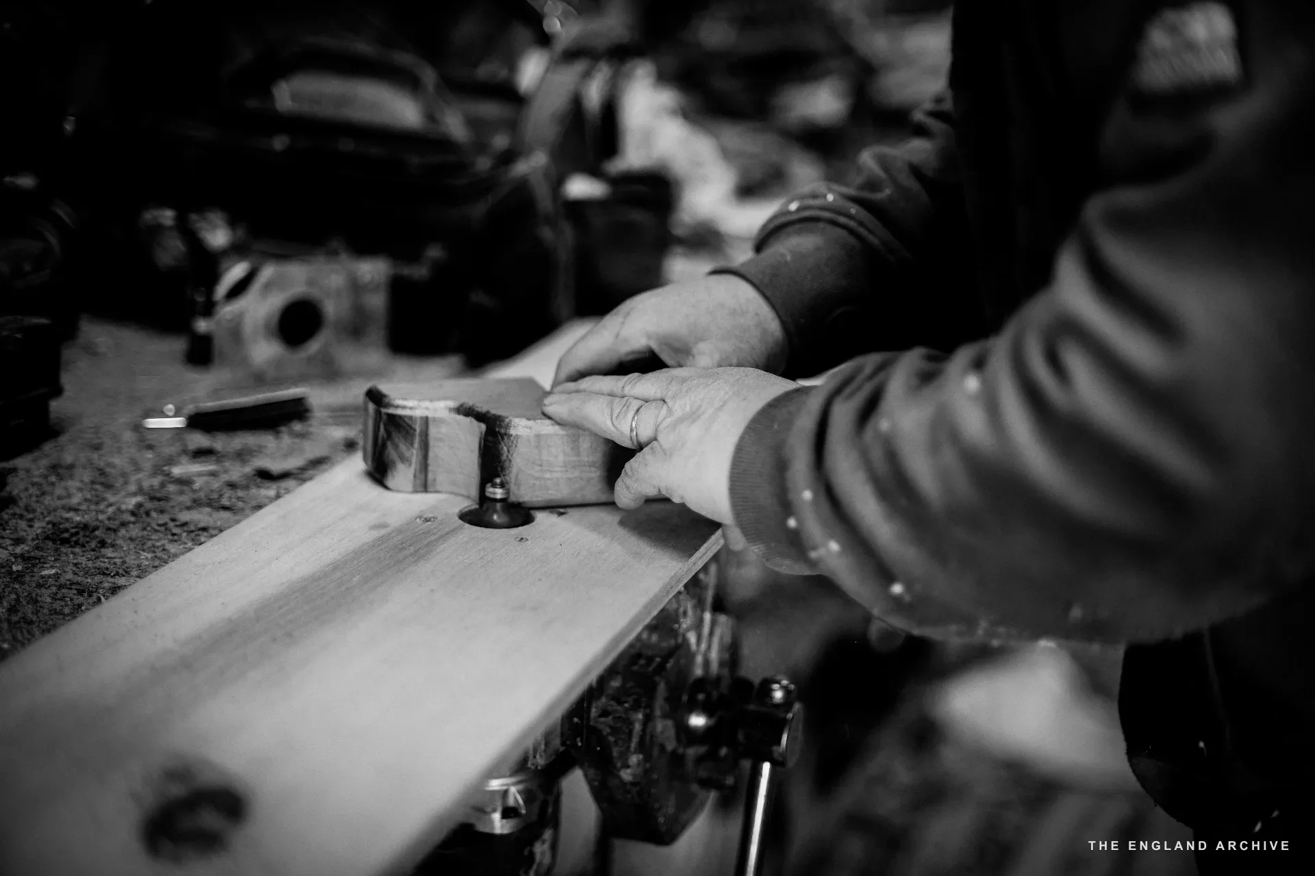 A close on two pairs of hands at the bench - one pair guiding a router along the edge of a curved timber piece, the other pair holding the workpiece steady. A wedding ring visible.