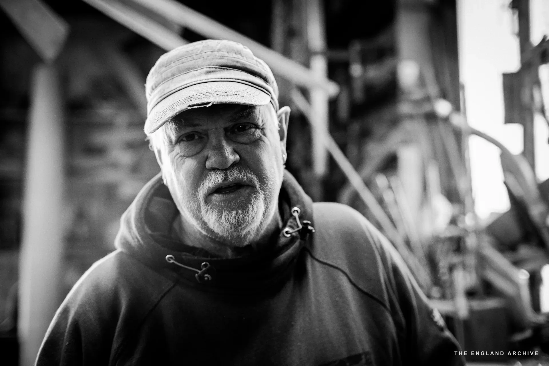 A head-and-shoulders portrait of a yard worker in a leather brimmed hat and grey beard, looking past the camera, the workshop dim behind him.
