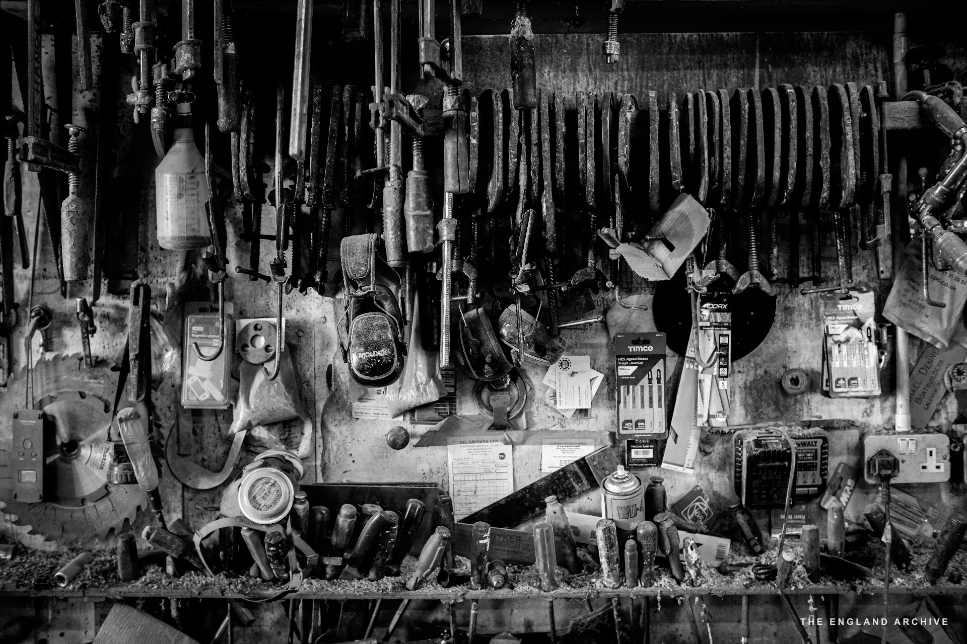 A close on the workshop tool wall: rows of hanging hand-tools - chisels, planes, callipers, gauges, pliers - against a dark backdrop, the bench surface in the foreground covered in shavings and small parts.