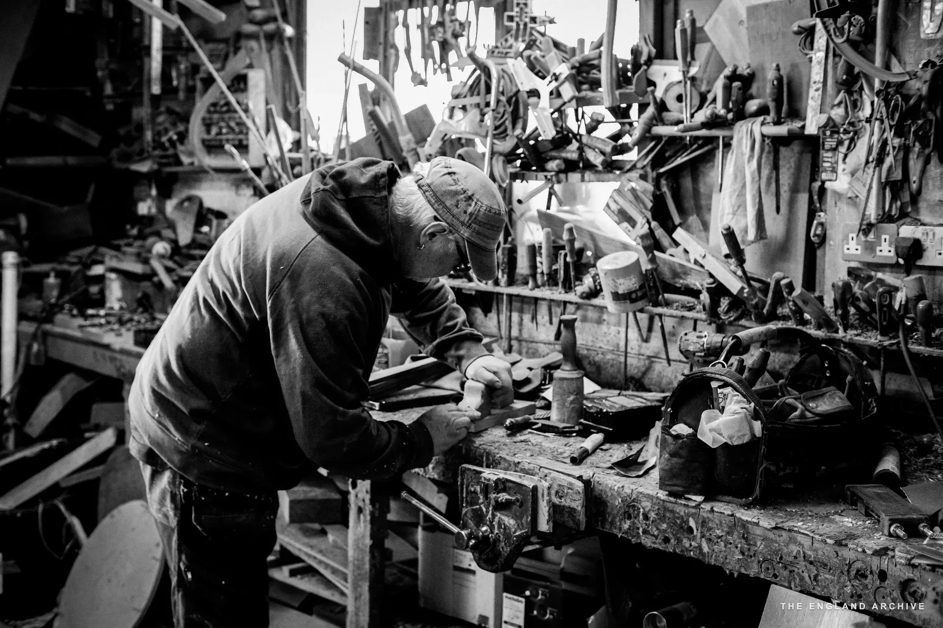A worker in a cap working alone at the tool wall bench, head down on a small workpiece, the dense rack of tools rising above and the workshop receding behind.