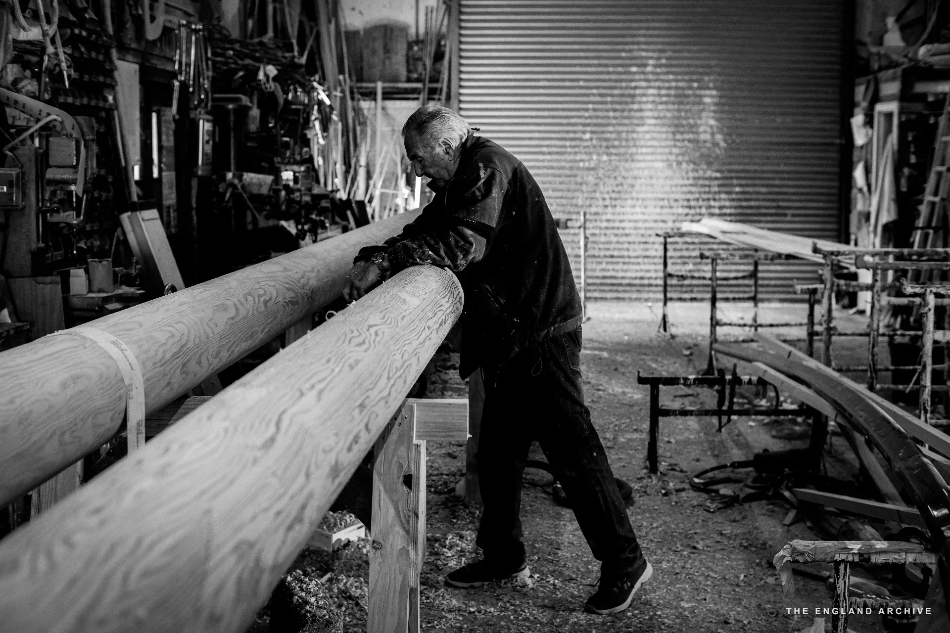 Michael Dennett working two long planks of timber at the bench, sleeves rolled, the workshop door behind him.