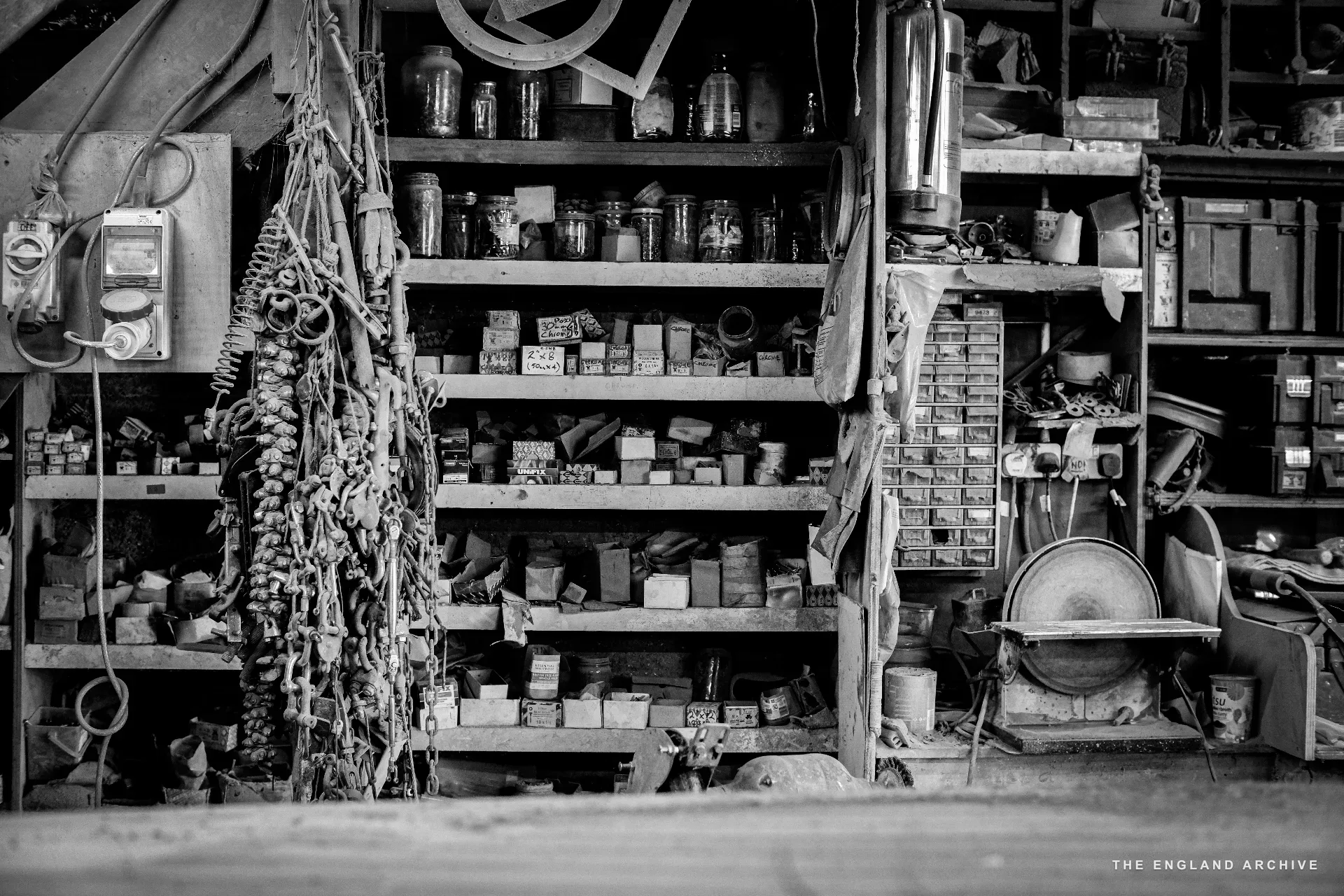A wide view of the workshop’s parts shelves - rows of small drawers and jars, hanging tools and ropes on the left, a circular saw blade and grinder on the right.