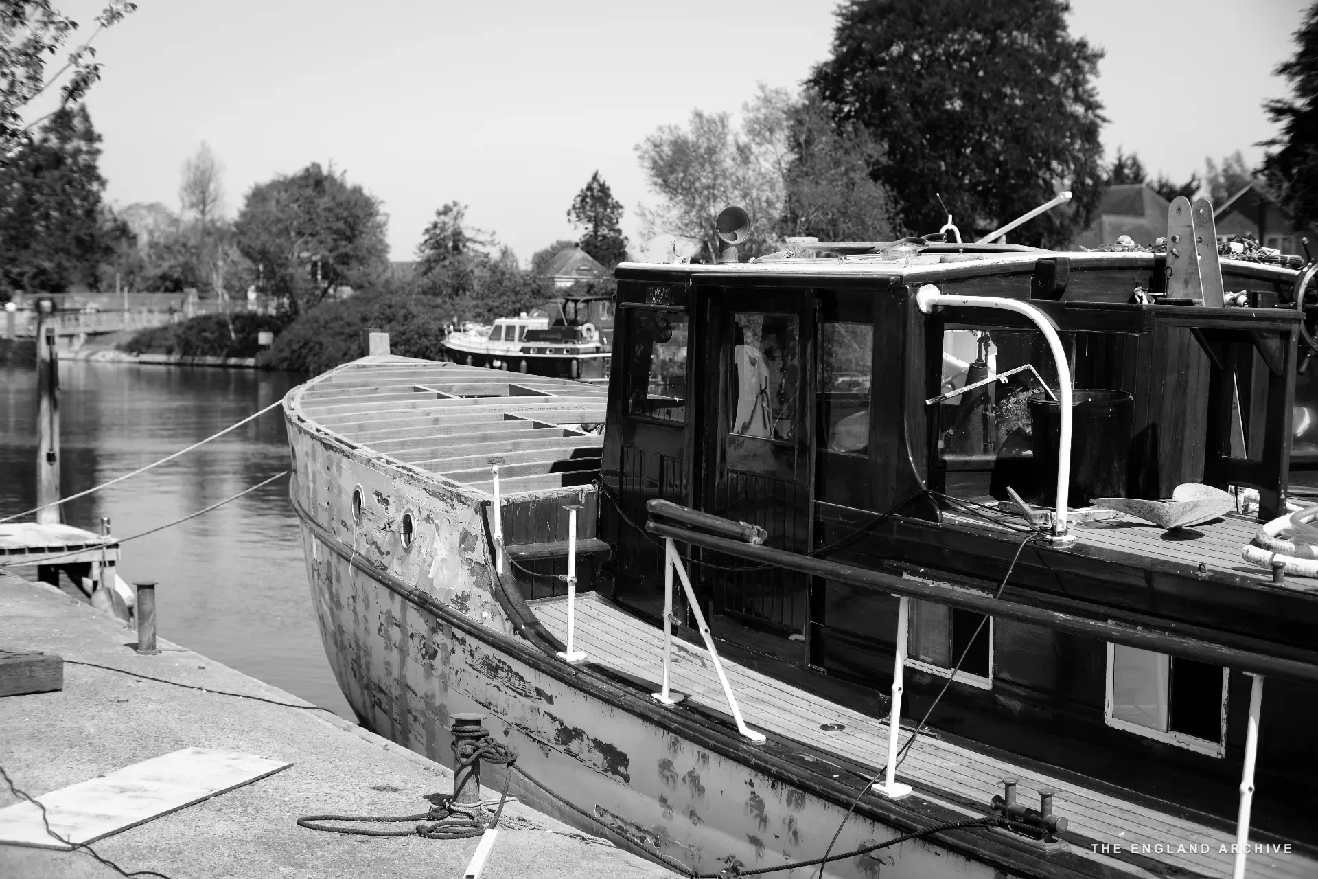 A long-cabin wooden river launch moored at the yard’s slipway, the river behind it and the willow-lined far bank, late-morning sun.