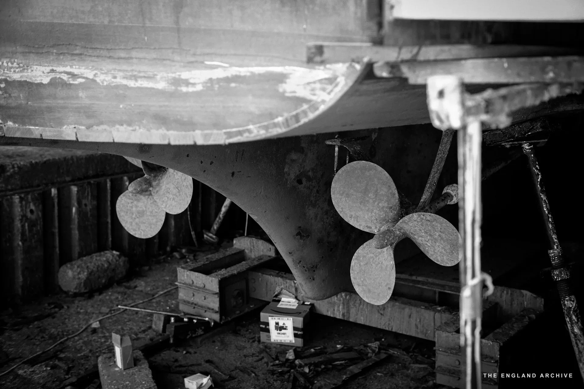 Twin propellers under the keel of a wooden hull in the workshop dry dock, the rudder between them, timber bracing of the cradle below and the boat’s underside above.