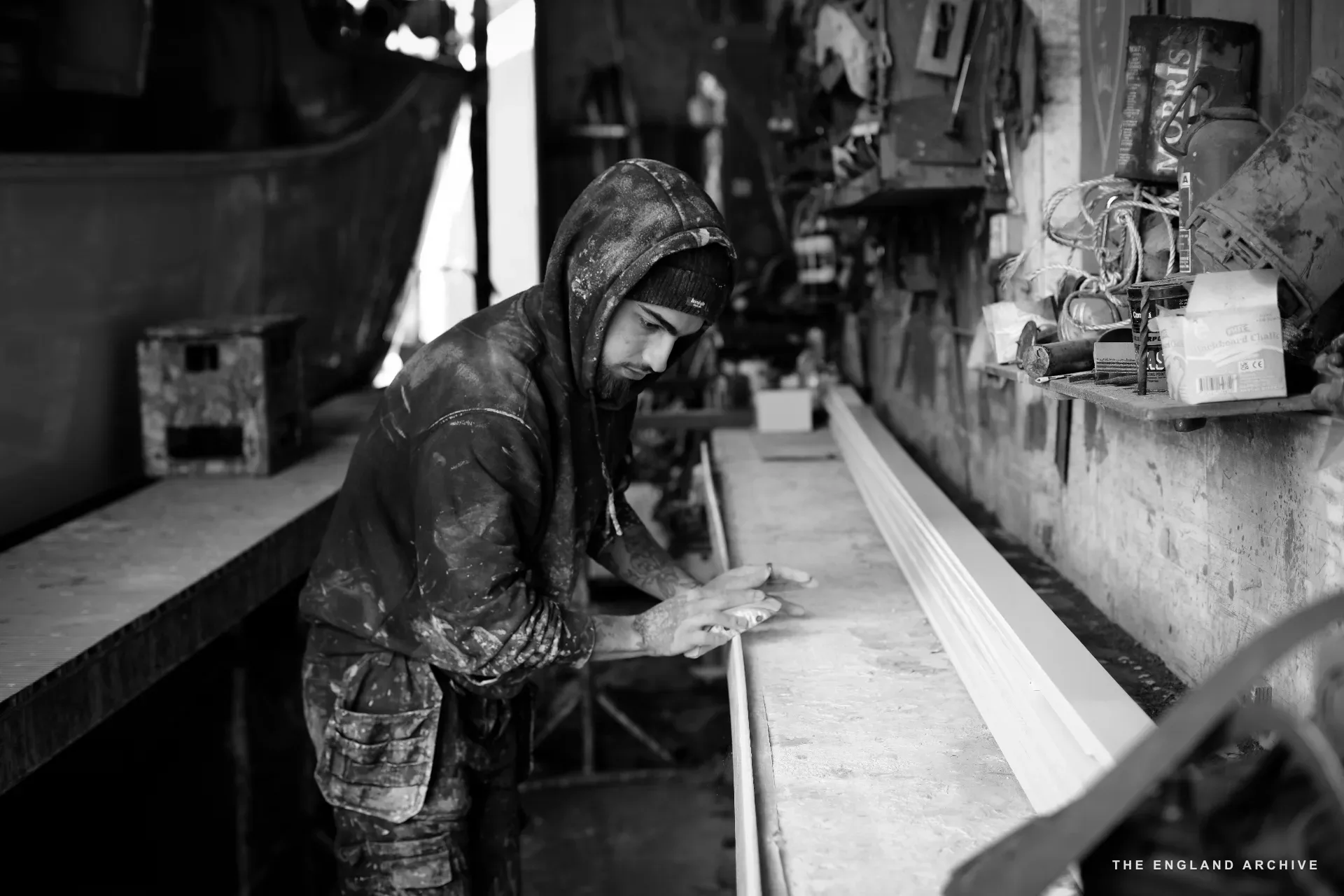 A young apprentice at the bench in the workshop - hooded sweatshirt covered in dust and resin, beanie hat under the hood, working a long timber along its edge with both hands. A row of tools and supplies on the shelf behind him.