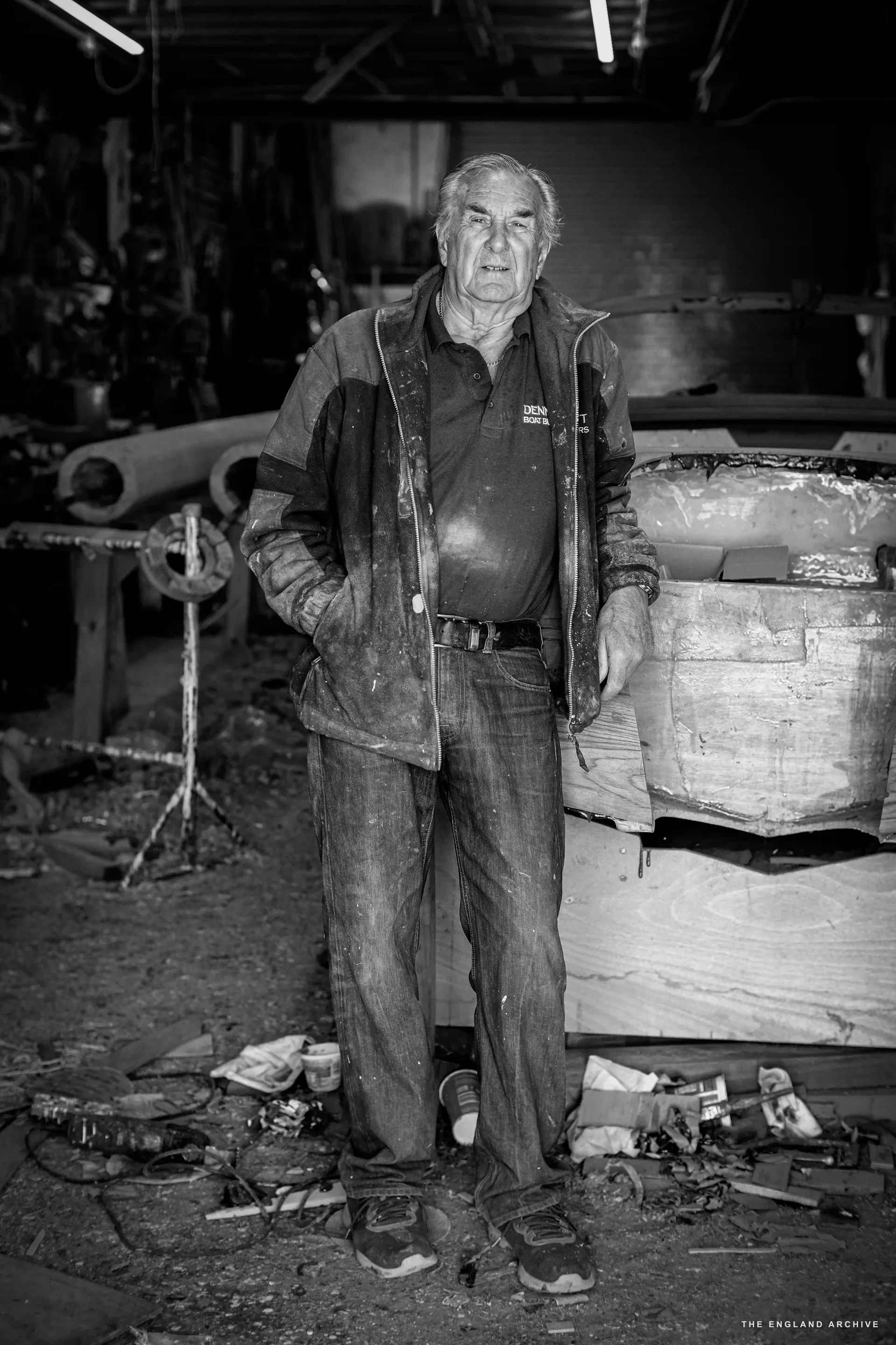 A formal full-length portrait of Michael Dennett standing in the workshop, hands in his pockets, tools and a partially built hull behind him, the workshop floor scattered with timber offcuts.