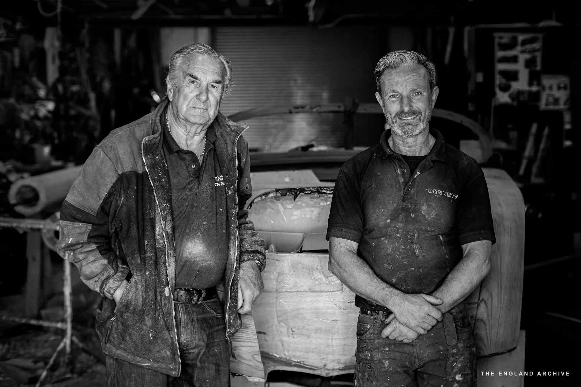 Joint portrait of Michael Dennett (left, in jacket) and Stephen Dennett (right, in DENNETT polo) standing side by side in the workshop, a partially built hull behind them, both looking directly at the camera.