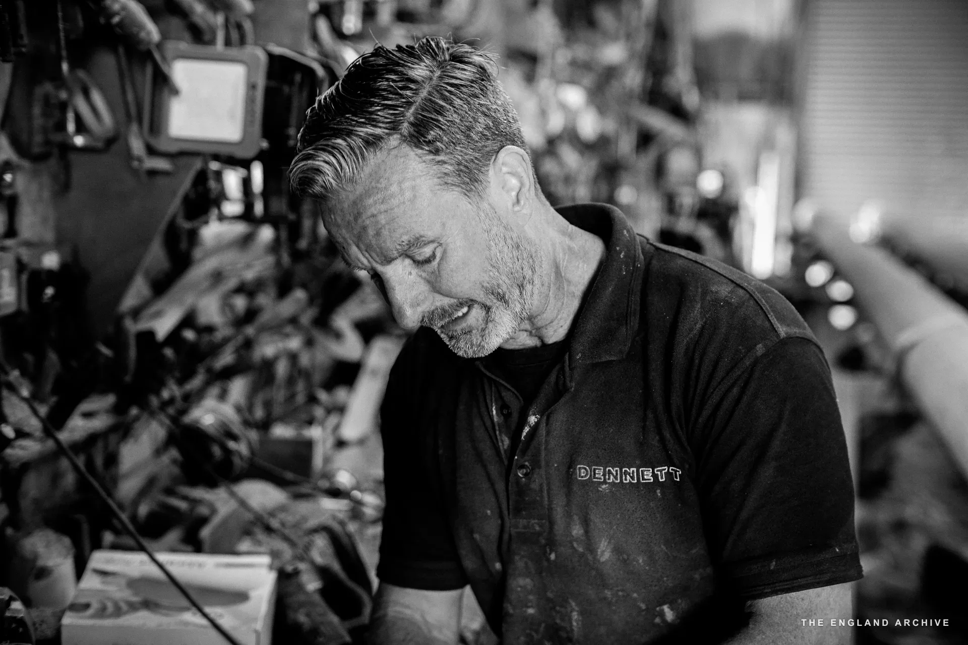 Stephen Dennett at the workbench in close half-light, head down on the work, the DENNETT polo and tool wall in shallow focus behind.