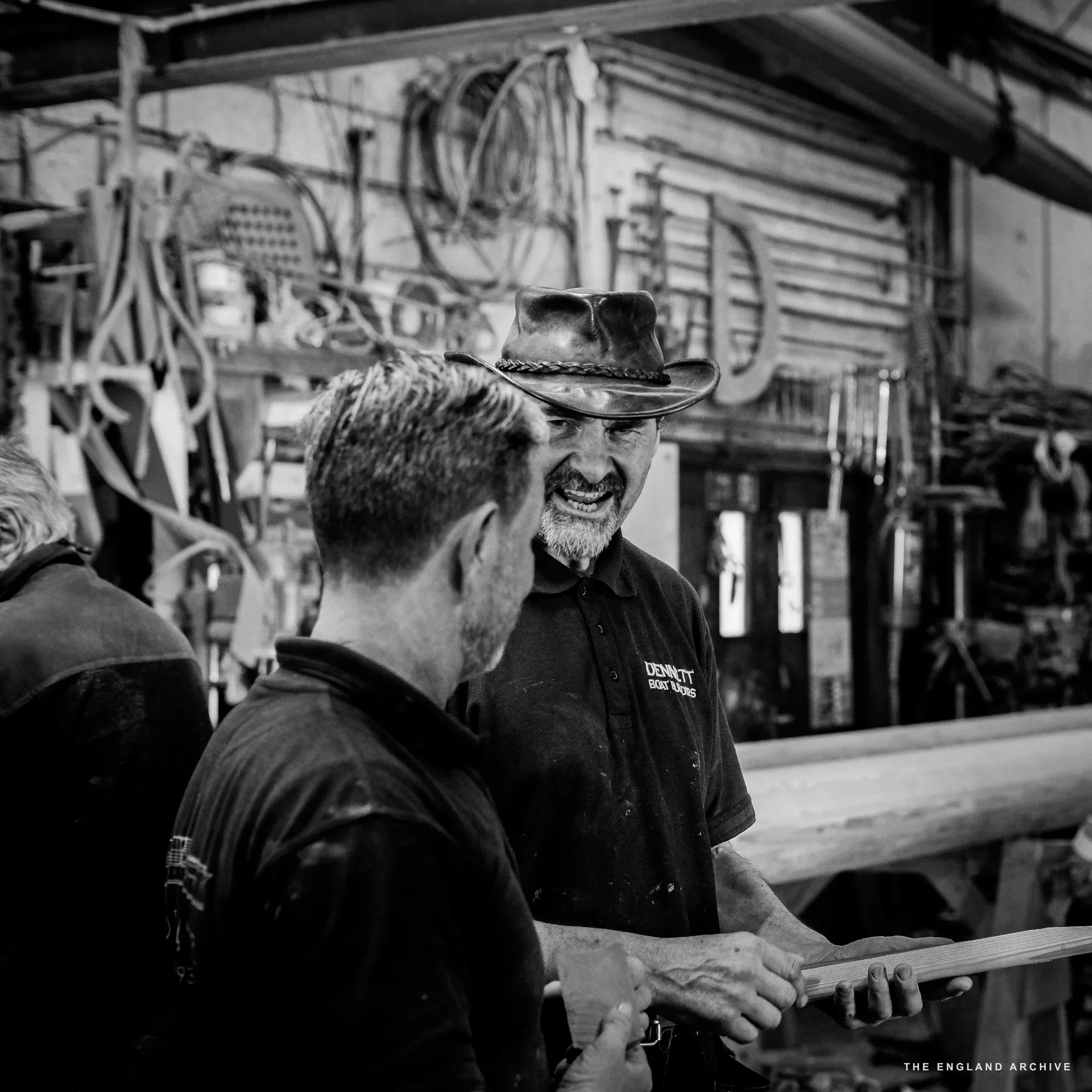 Stephen Dennett (foreground left, back to camera) in conversation with the leather-hatted yard worker (centre, smiling broadly through his grey beard), the workshop’s tool wall behind them.