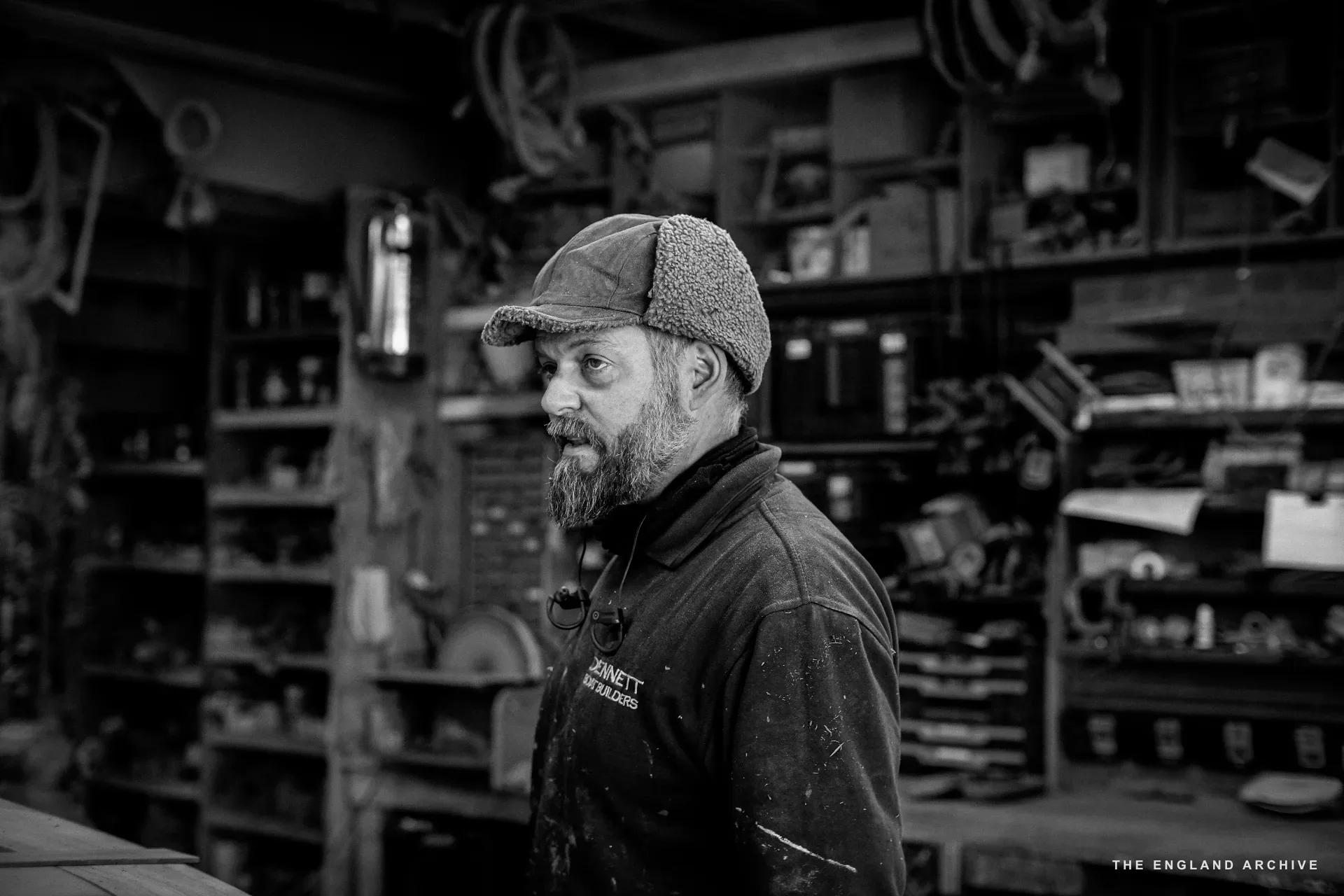 A worker in a fur-lined cap and grey beard standing at the workbench, looking past the camera, the workshop’s parts shelves and the cluttered bench behind him.