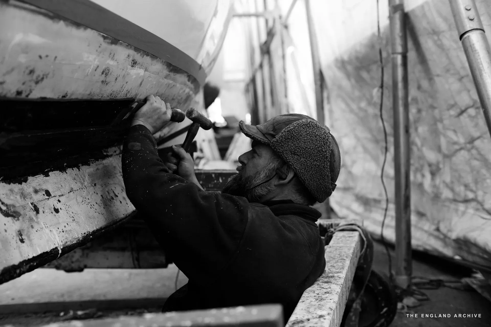 A worker in a knitted cap reaching up under the side of a hull at the workshop, working at the planking from underneath, the boat’s curve filling the upper half of the frame.