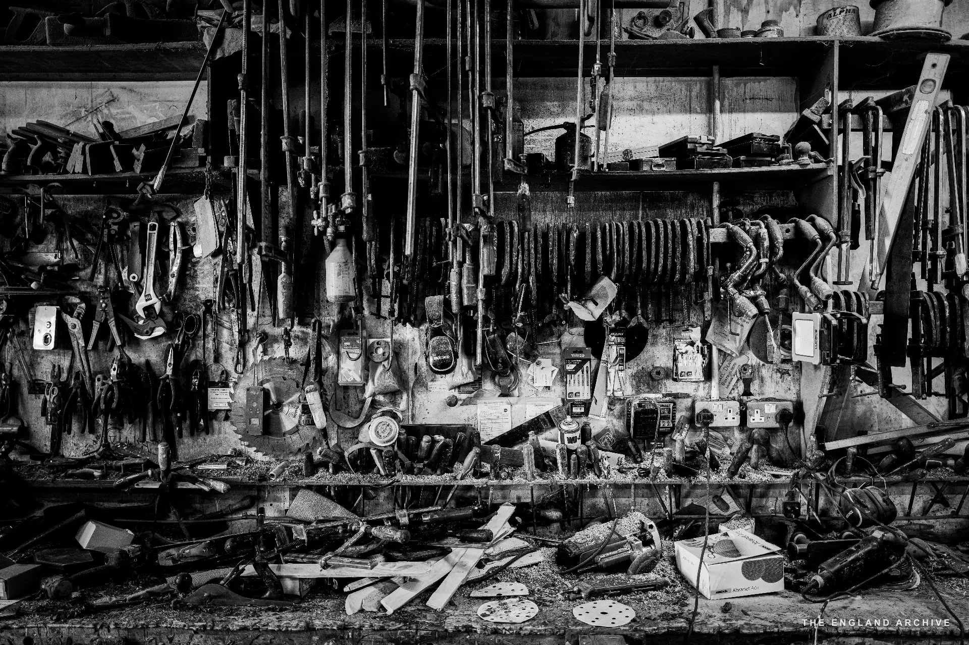 A wide view of the workshop’s tool wall and bench - hundreds of hand tools hung in racks above a long bench cluttered with parts, jars and offcuts. The bench surface in the foreground is heavily worked.